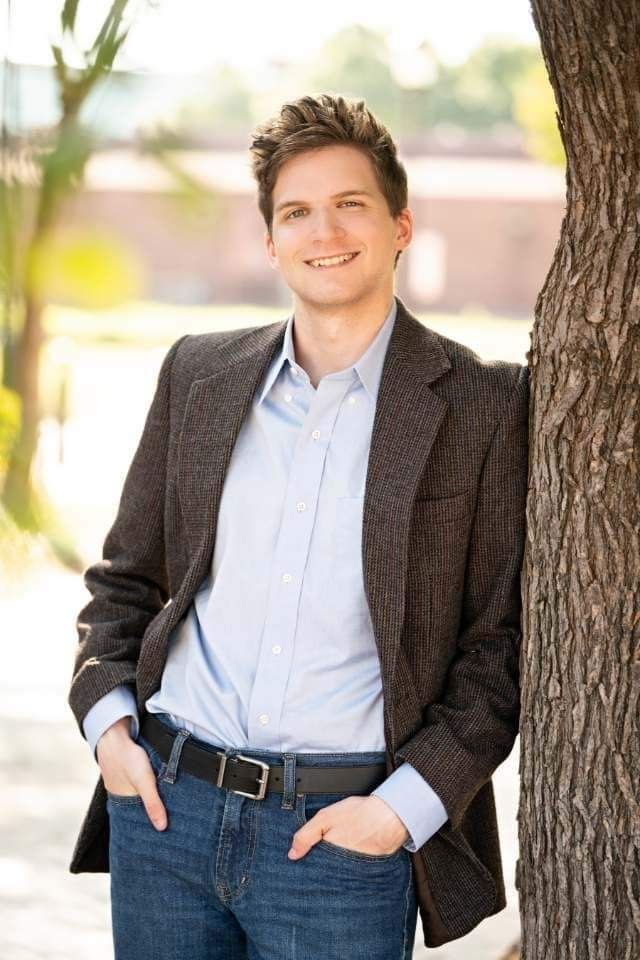 A young man with light skin, brown hair, and a friendly smile leaning against a tree outdoors, wearing a grey blazer, light blue button-up shirt, and jeans, with hands in his pockets.