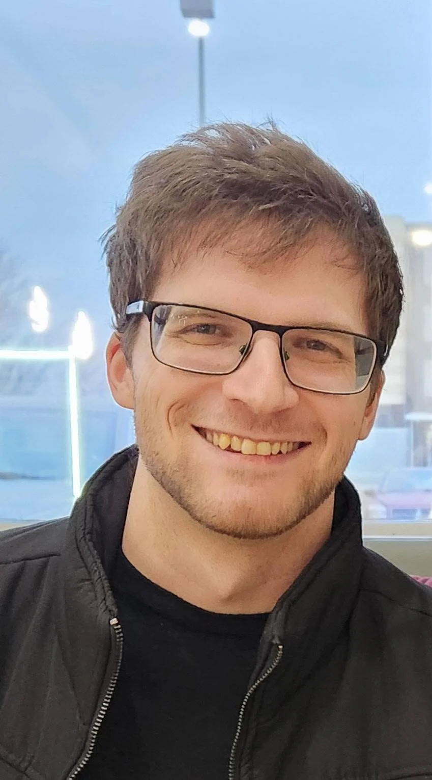 Portrait of a smiling young man wearing glasses, black jacket, and black shirt, indoors with a window behind, showing blue sky and outdoor elements.