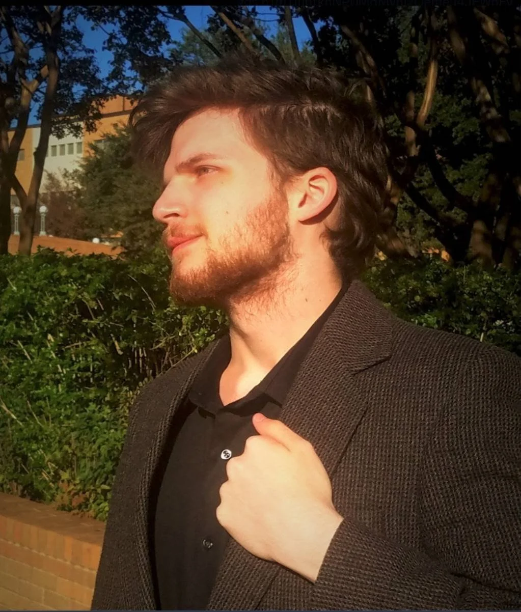 A young man with brown hair and a beard, wearing a dark blazer and black shirt, is standing outdoors in front of trees and bushes during sunset.