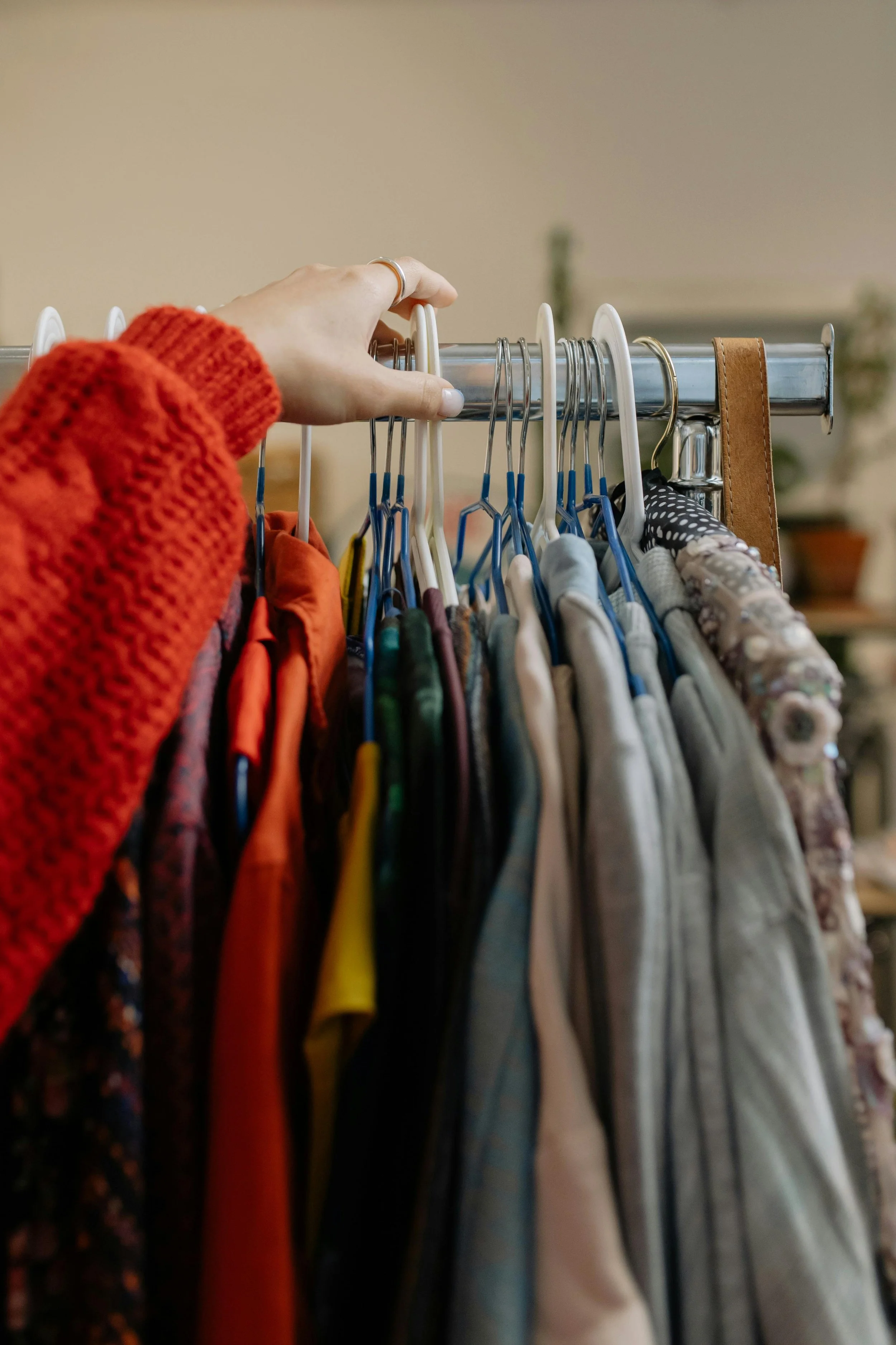 A hand with a silver ring reaching for a clothing hanger with various colorful outfits hanging on a metal rack.