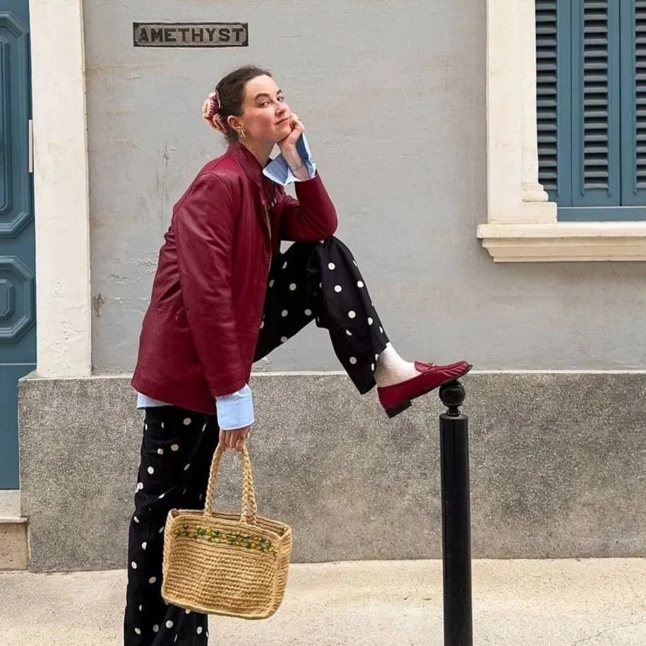 A woman with a red jacket, polka dot pants, and red shoes leaning on a black post, with her foot on top of it. She is holding a woven basket and standing in front of a building with blue shutters. There is a sign that reads 'AMETHYST' on the wall behind her.