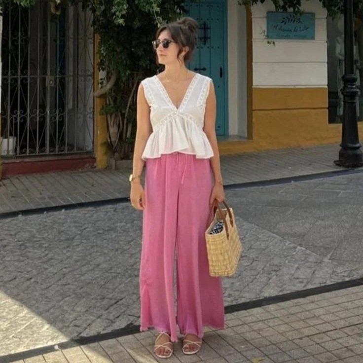 A woman in a white sleeveless top and pink wide-leg pants, wearing sunglasses and holding a woven straw bag, standing on a city sidewalk.