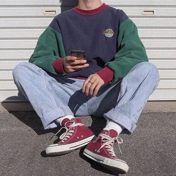 Person sitting cross-legged outdoors, wearing a colorful sweatshirt, light-wash jeans, and red sneakers, holding a smartphone.