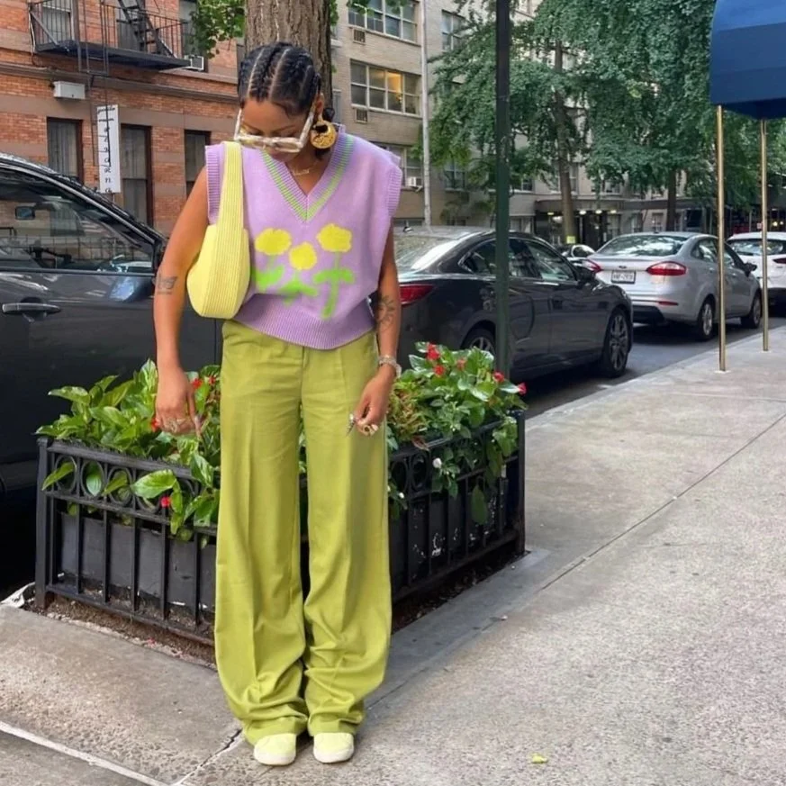 A woman standing on a city sidewalk wearing a lavender sleeveless sweater vest with yellow flower designs, lime green wide-leg pants, and white shoes. She has shoulder-length braided hair, large sunglasses, and carries a yellow bag.