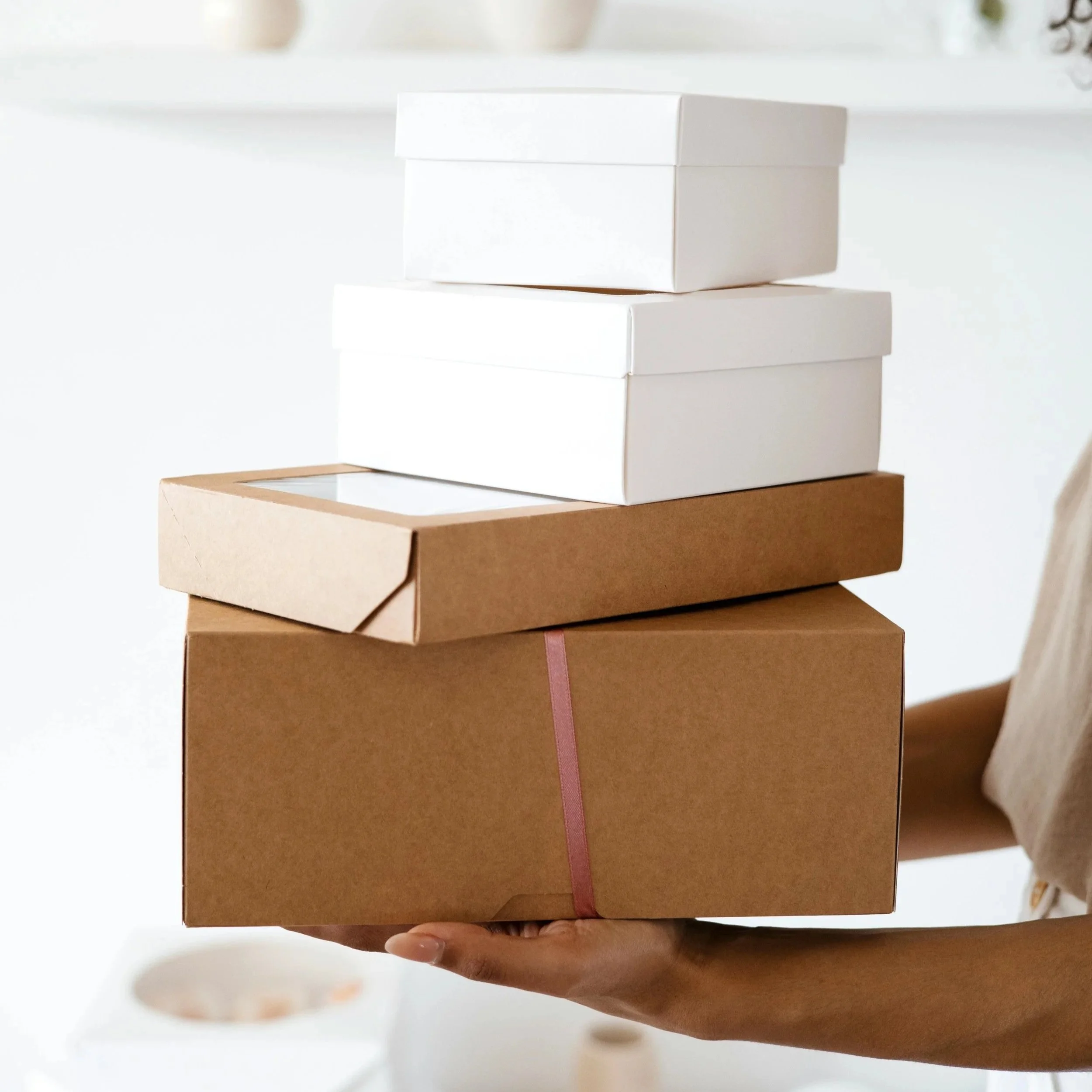 Person holding stacked boxes of varying sizes and colors, including brown, white, and cardboard, with one hand visible at the bottom of the stack.
