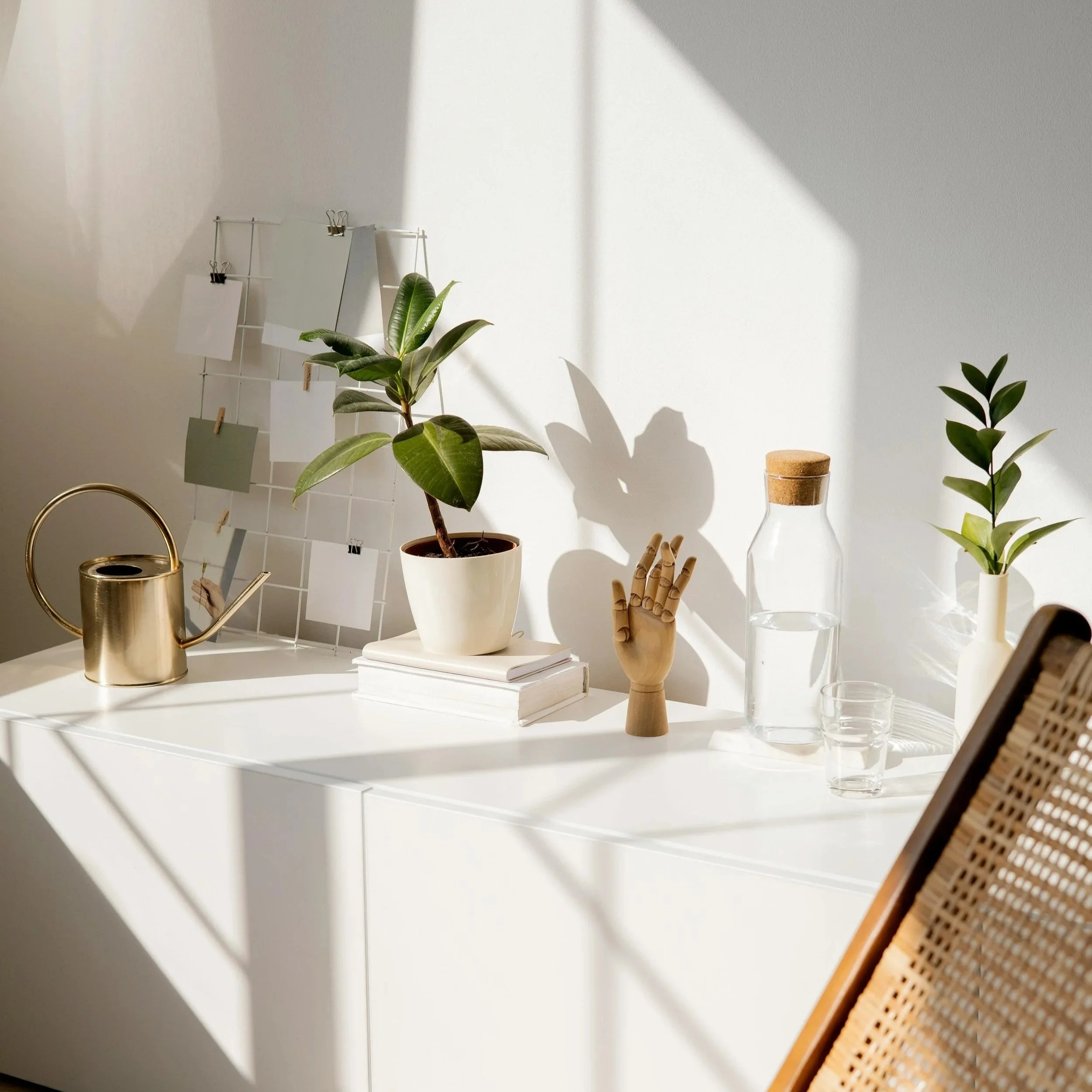 A minimalist workspace with a white desk, green potted plants, a gold watering can, a clear glass, and a woven chair, illuminated by natural sunlight.