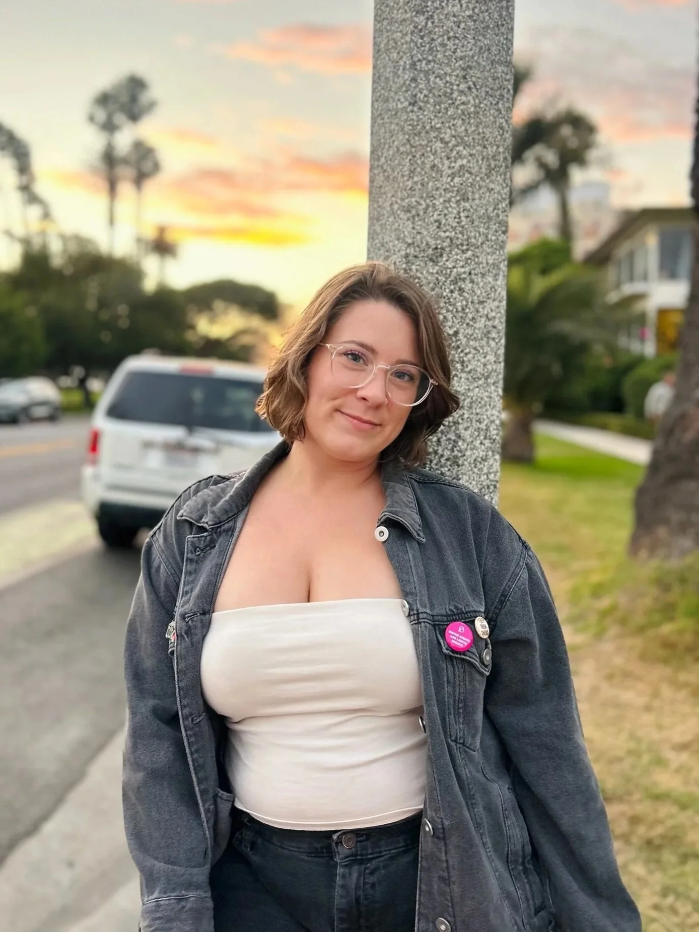 A woman with short brown hair, glasses, and a black denim jacket, smiling while standing outside near a gray street lamp post during sunset, with trees, houses, and parked cars in the background.