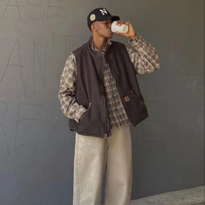 A young person drinking from a paper cup while standing against a gray wall with faint writing.