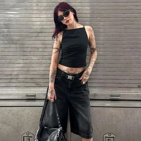 Woman with dark hair and sunglasses standing against a gray, horizontal wooden wall, wearing a black sleeveless top, black shorts, and holding a black bag.
