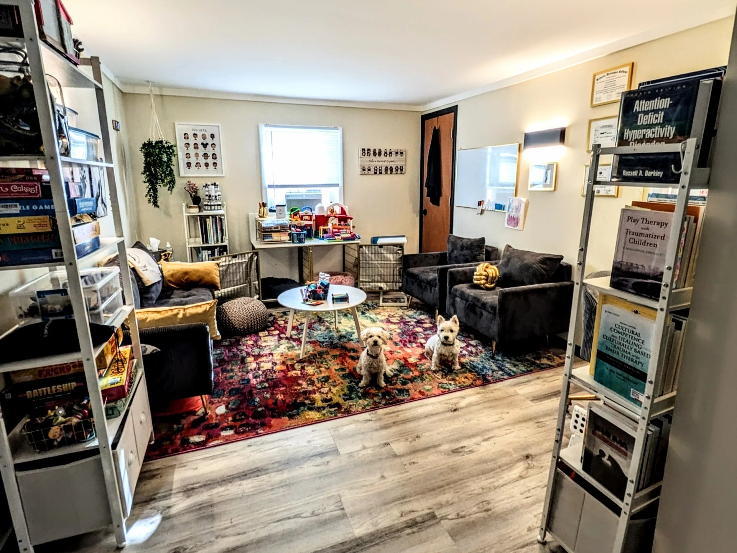 A cozy room with bookshelves, a colorful rug, and a sitting area with two small dogs. The room has a window, a whiteboard, and framed artwork on the walls. There are tables with toys and books, and multiple chairs and couches for seating.