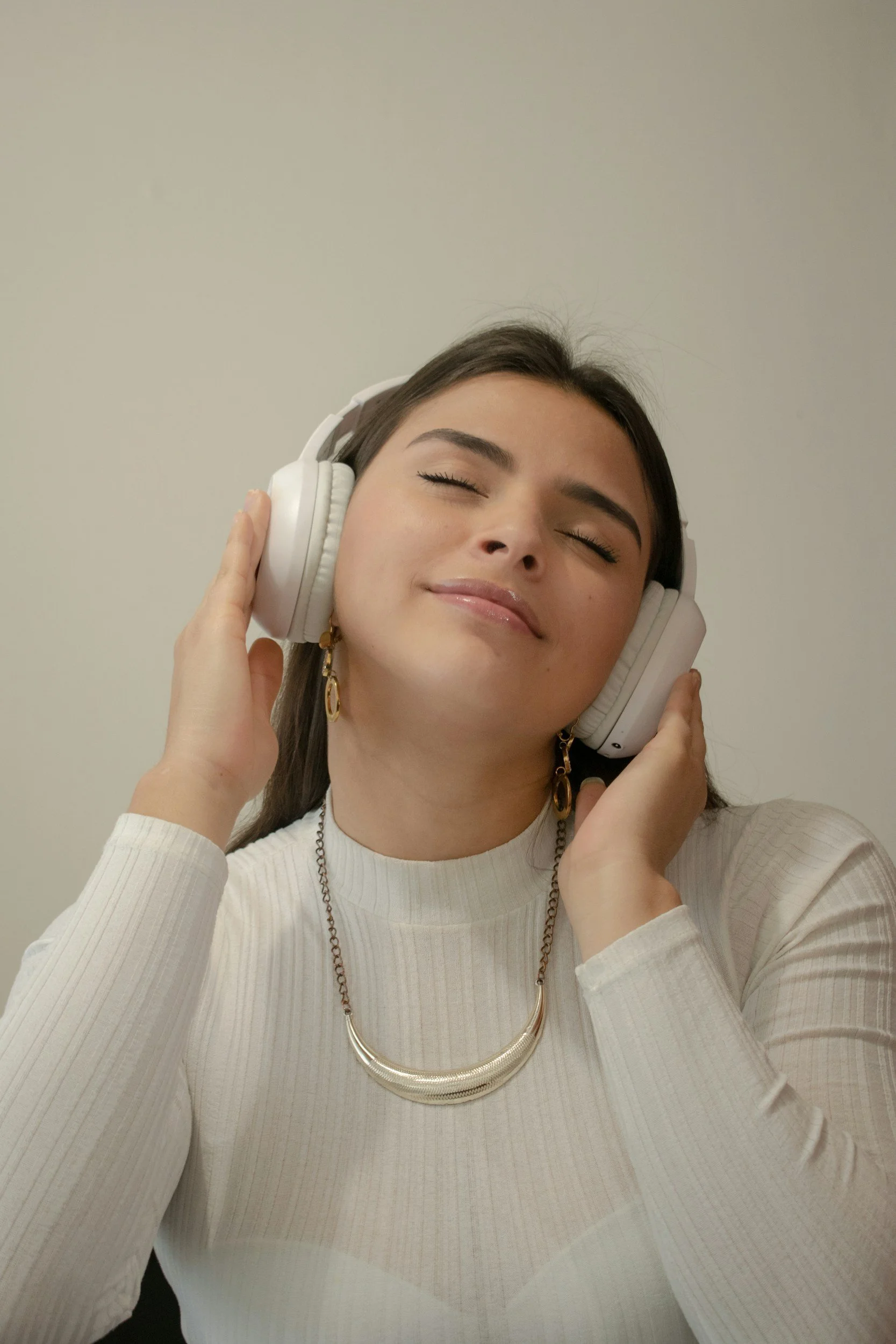 A woman with long dark hair, wearing a white ribbed long-sleeve top, gold earrings, and a gold necklace, smiling with eyes closed and listening to music with white headphones.