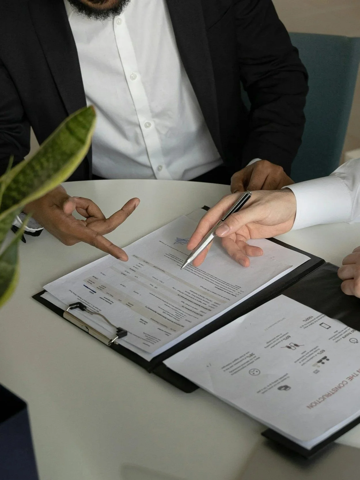 Two people reviewing and signing documents at a table, with a clipboard holding papers and a pen in one person's hand. A plant is partially visible on the left.