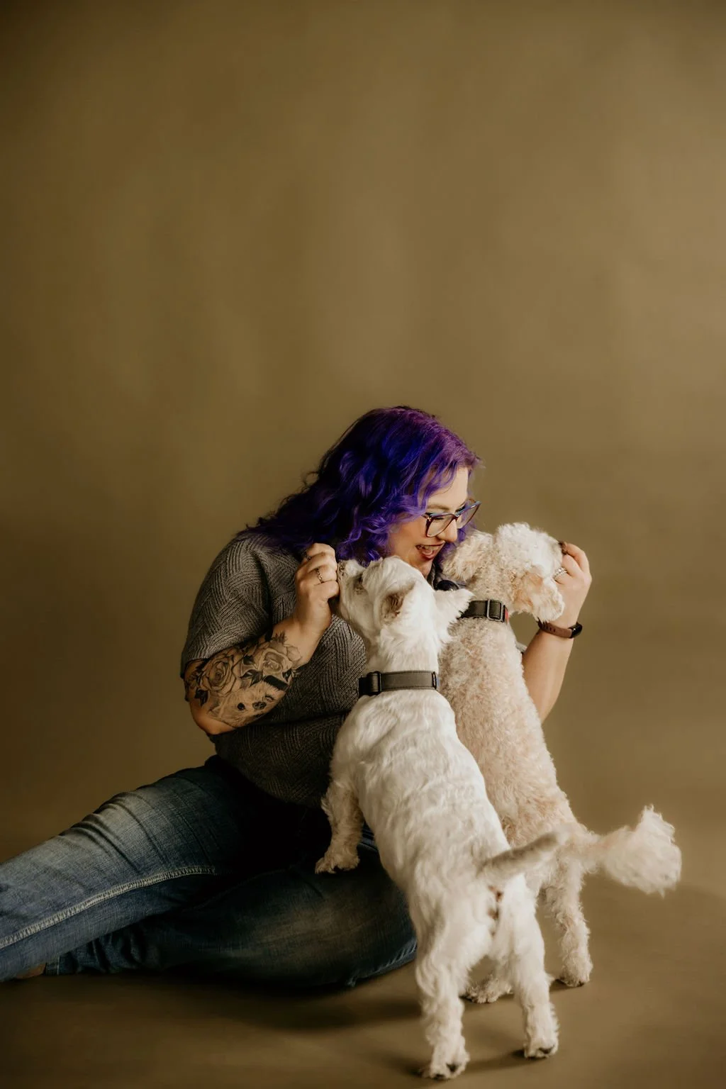 Woman with purple hair and glasses hugging three small dogs against a plain beige background.