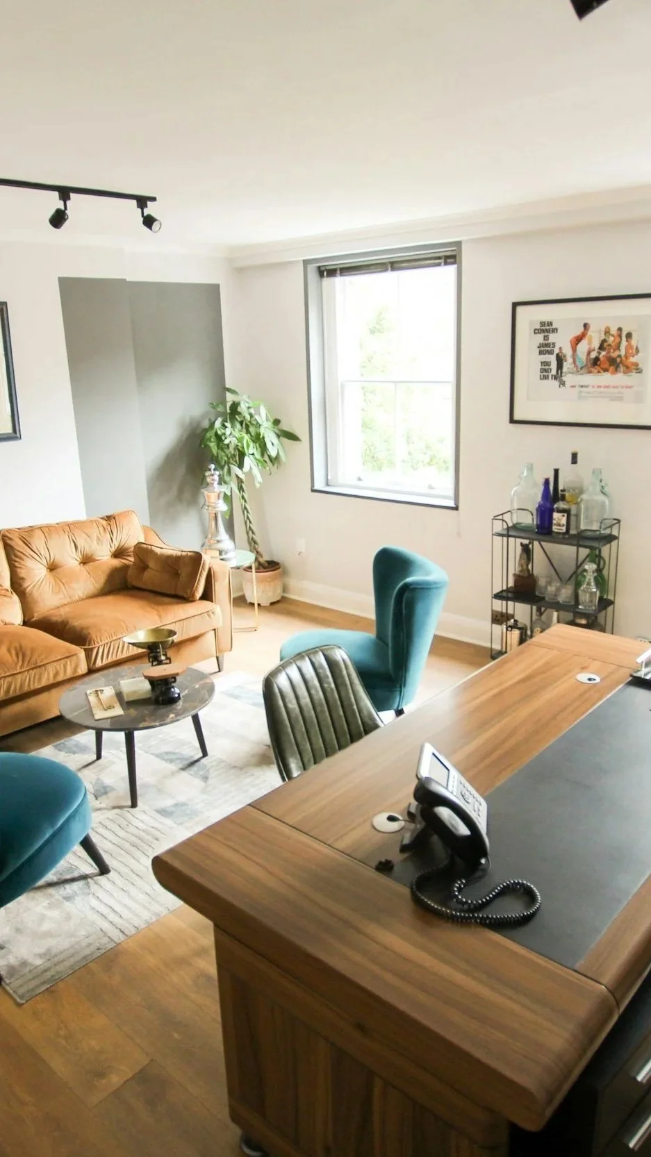 Office lounge with a leather sofa, teal chairs, a wooden desk, a bar cart with bottles, wall art, and a large window letting in natural light.