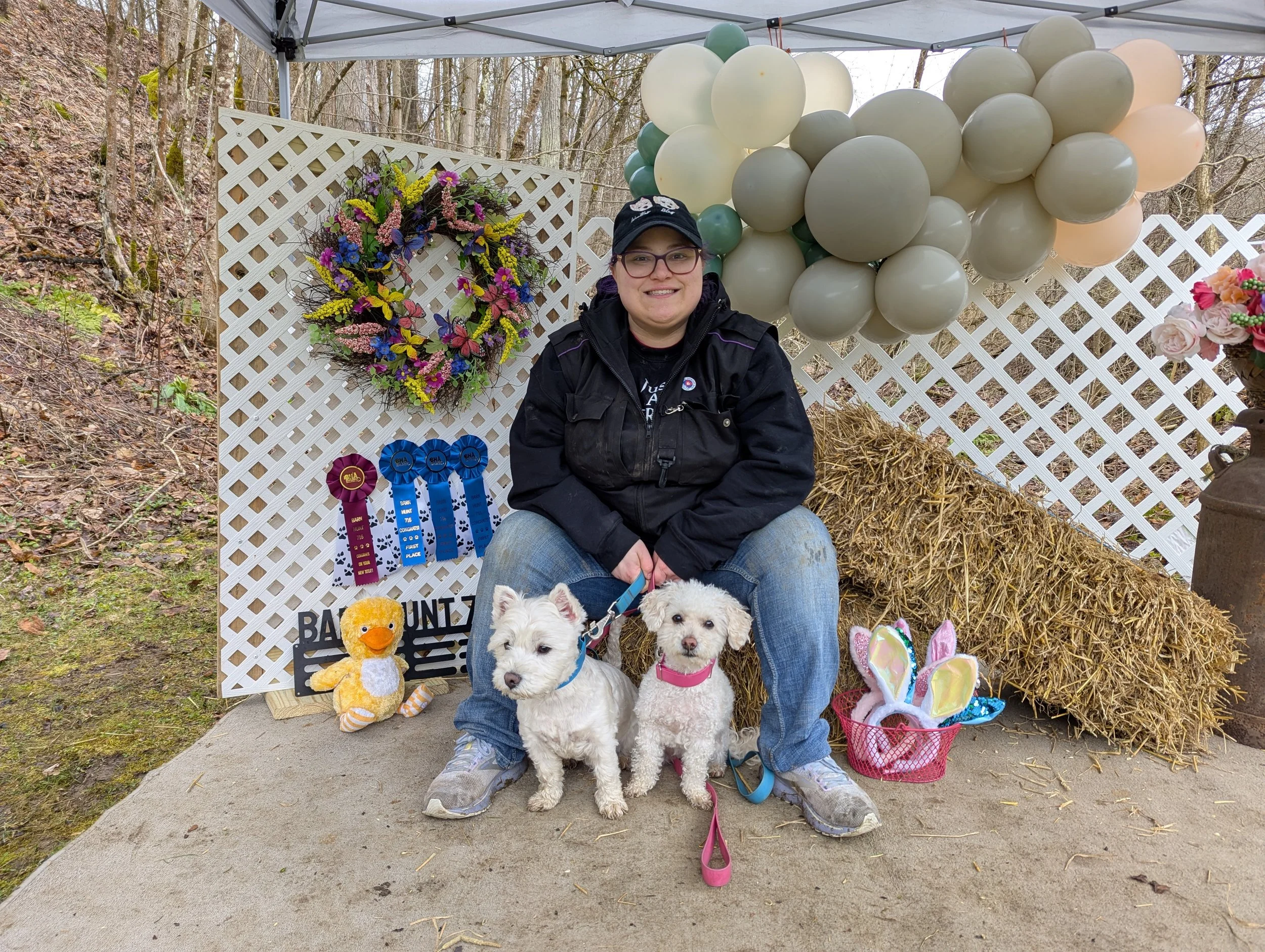 A woman sitting in front of a decorative display with balloons, a flower wreath, and ribbon awards, surrounded by stuffed animals and plush toys, in an outdoor wooded area during Easter or springtime celebration.