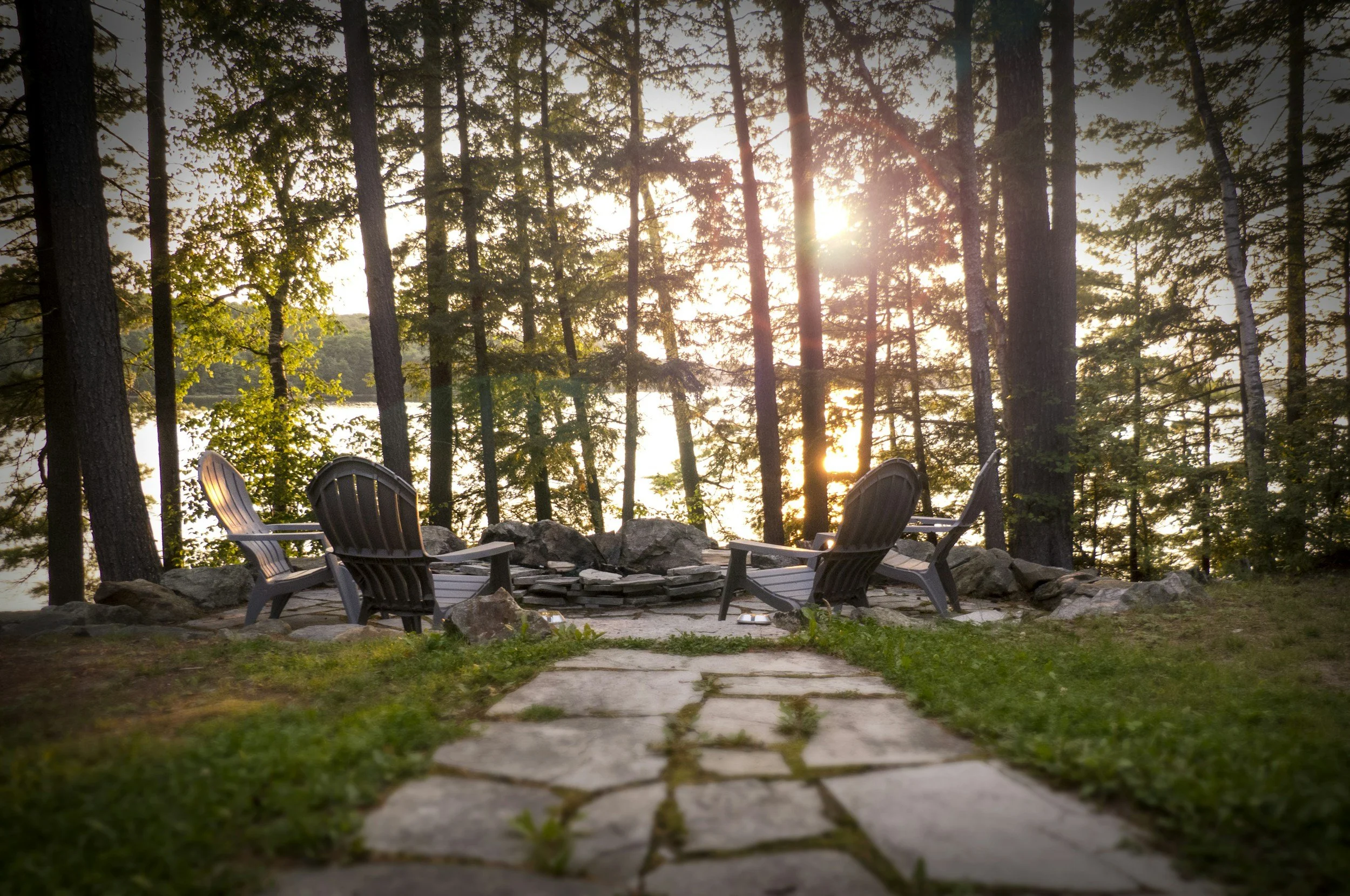 A sunset or sunrise view through a forest of tall trees with a lake visible in the background, and Adirondack chairs around a fire pit in the foreground.