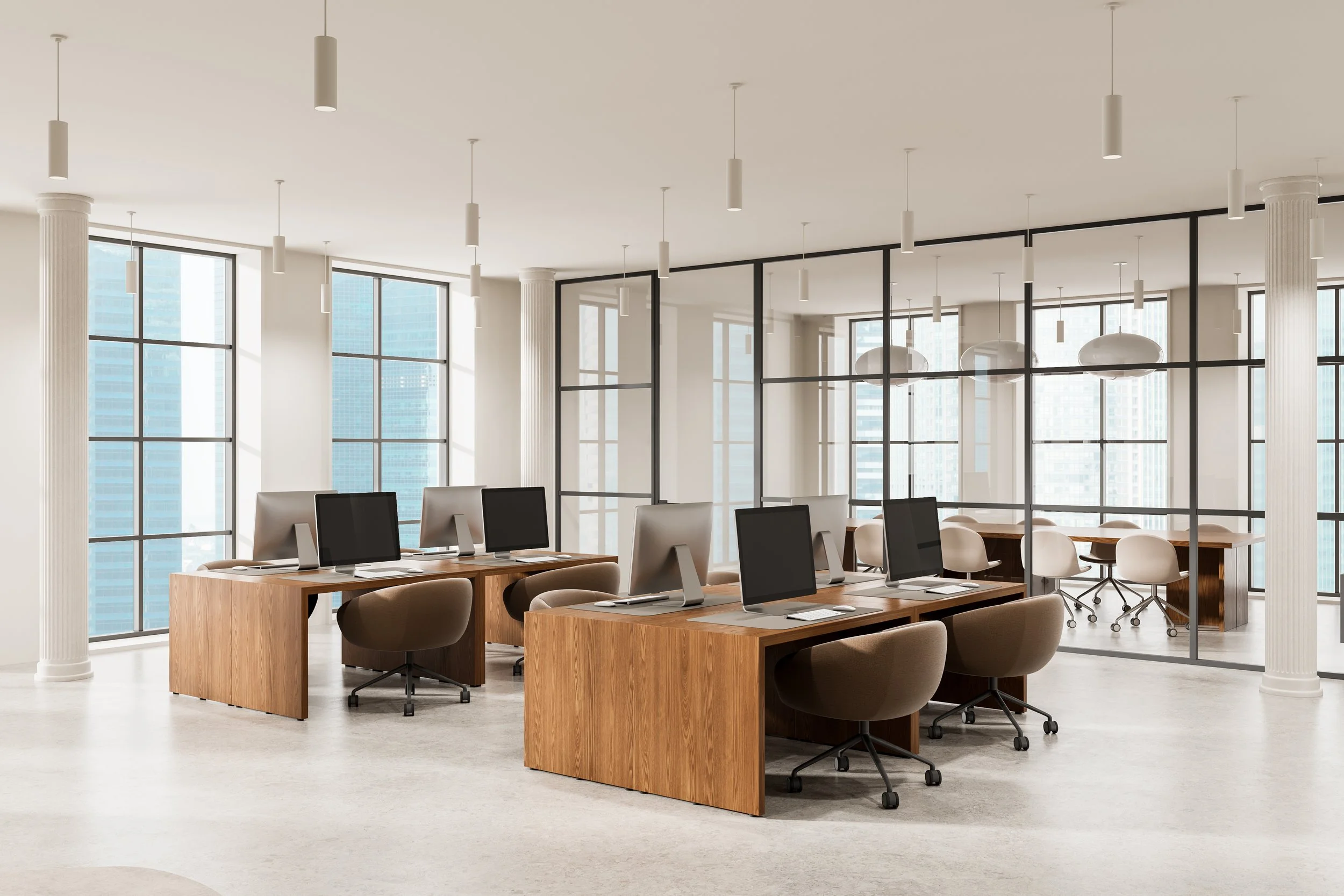 Modern office space with large windows, white walls, and high ceilings. Multiple desks with computers and chairs arranged in rows, with white and wooden furniture. Pendant lights hanging from the ceiling and large glass partitions separate different areas of the office.