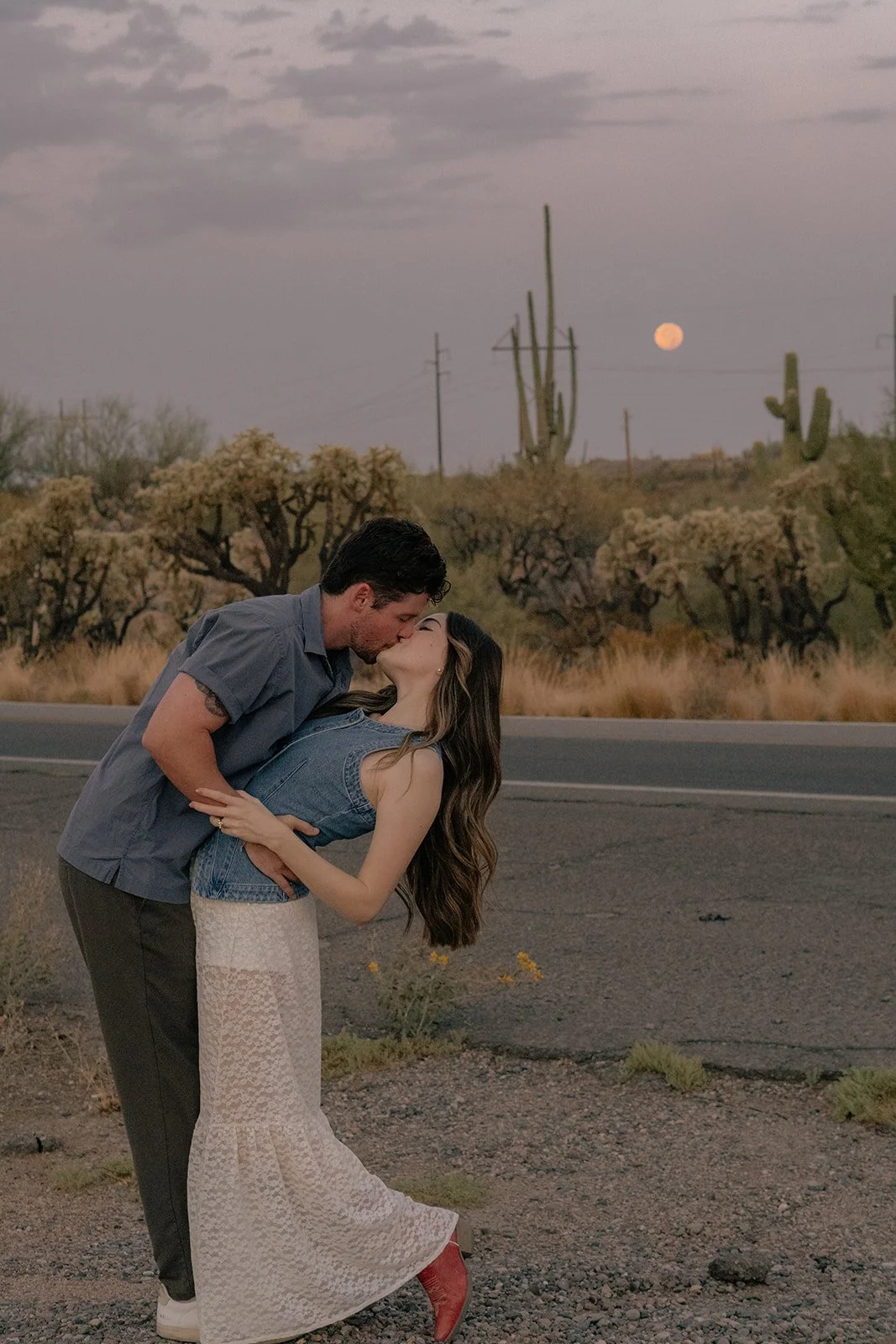 A couple sharing a kiss on a desert roadside during sunset with a full moon in the sky, and desert cacti and bushes in the background.