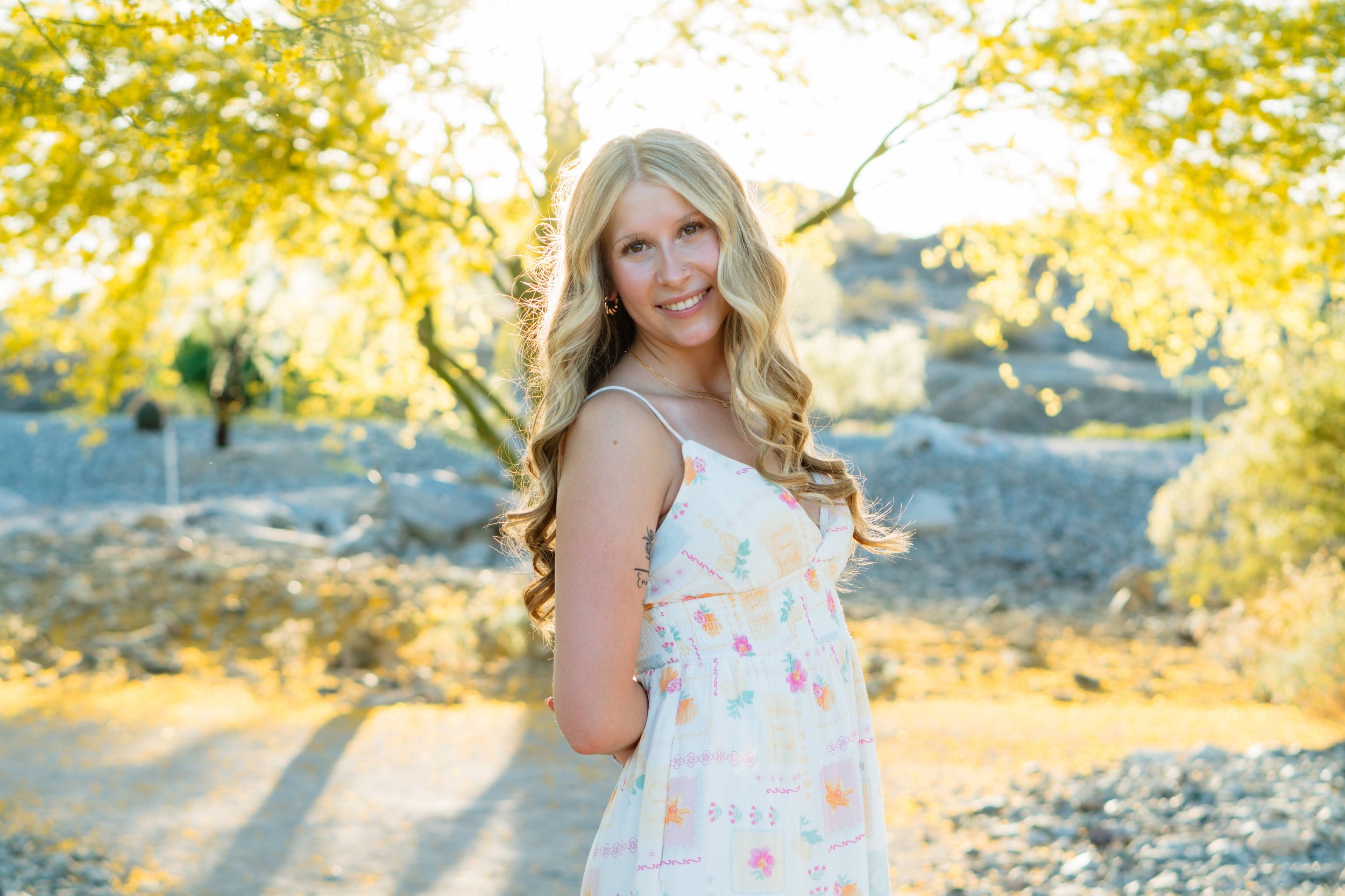 A young woman with long wavy blonde hair outdoors in spring, wearing a white floral dress, smiling at the camera with sunlight filtering through yellow-green trees in the background.