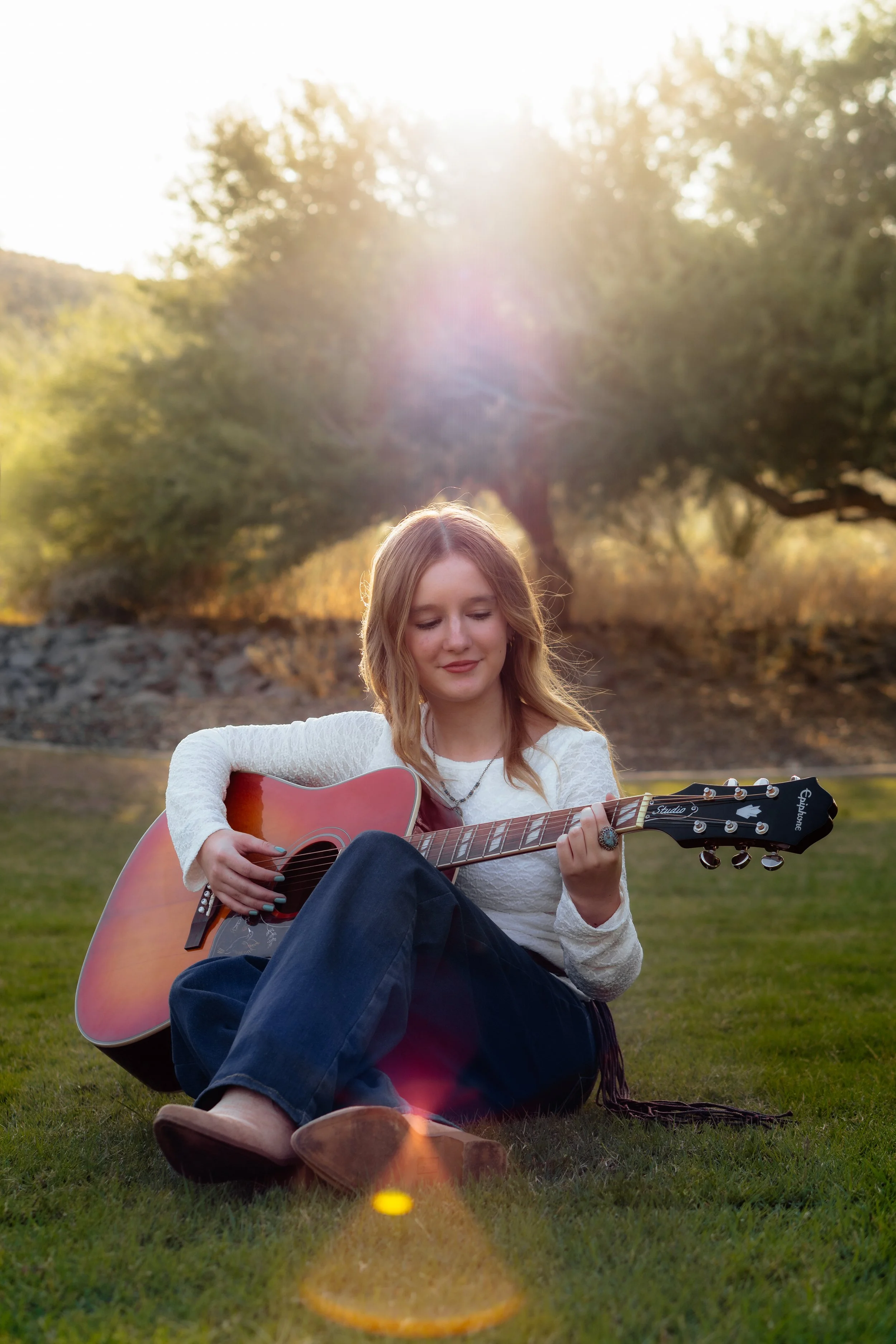 Young woman with long blonde hair playing an acoustic guitar outdoors on a grassy area during sunset with trees in the background.