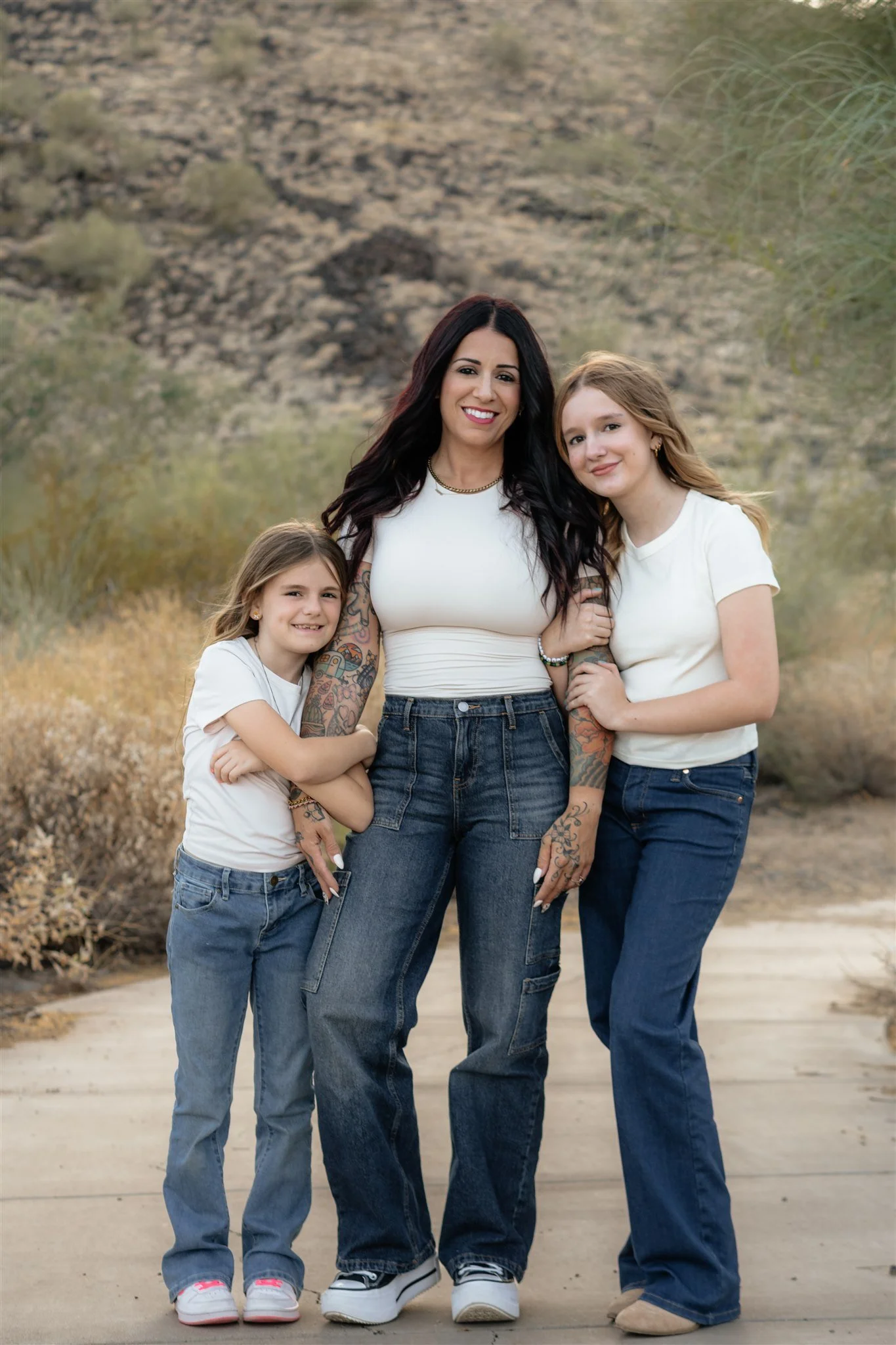A woman with black hair and two young girls pose outdoors in a desert landscape with hills and sparse vegetation, all dressed in casual white tops and blue jeans.