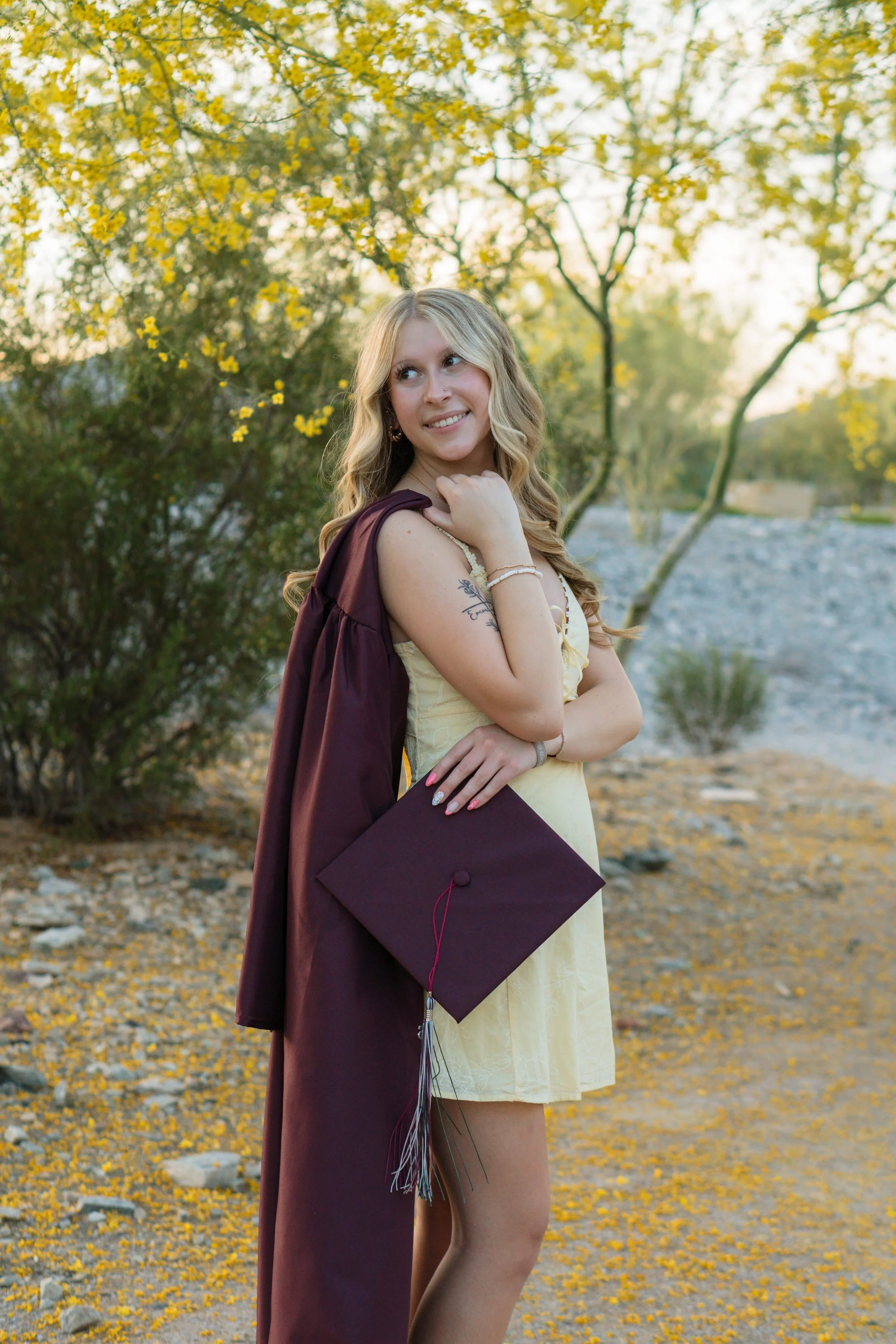 Young woman in a yellow dress holding a graduation cap, with a maroon sash, standing outdoors near a river with trees and yellow leaves in the background, during sunset.