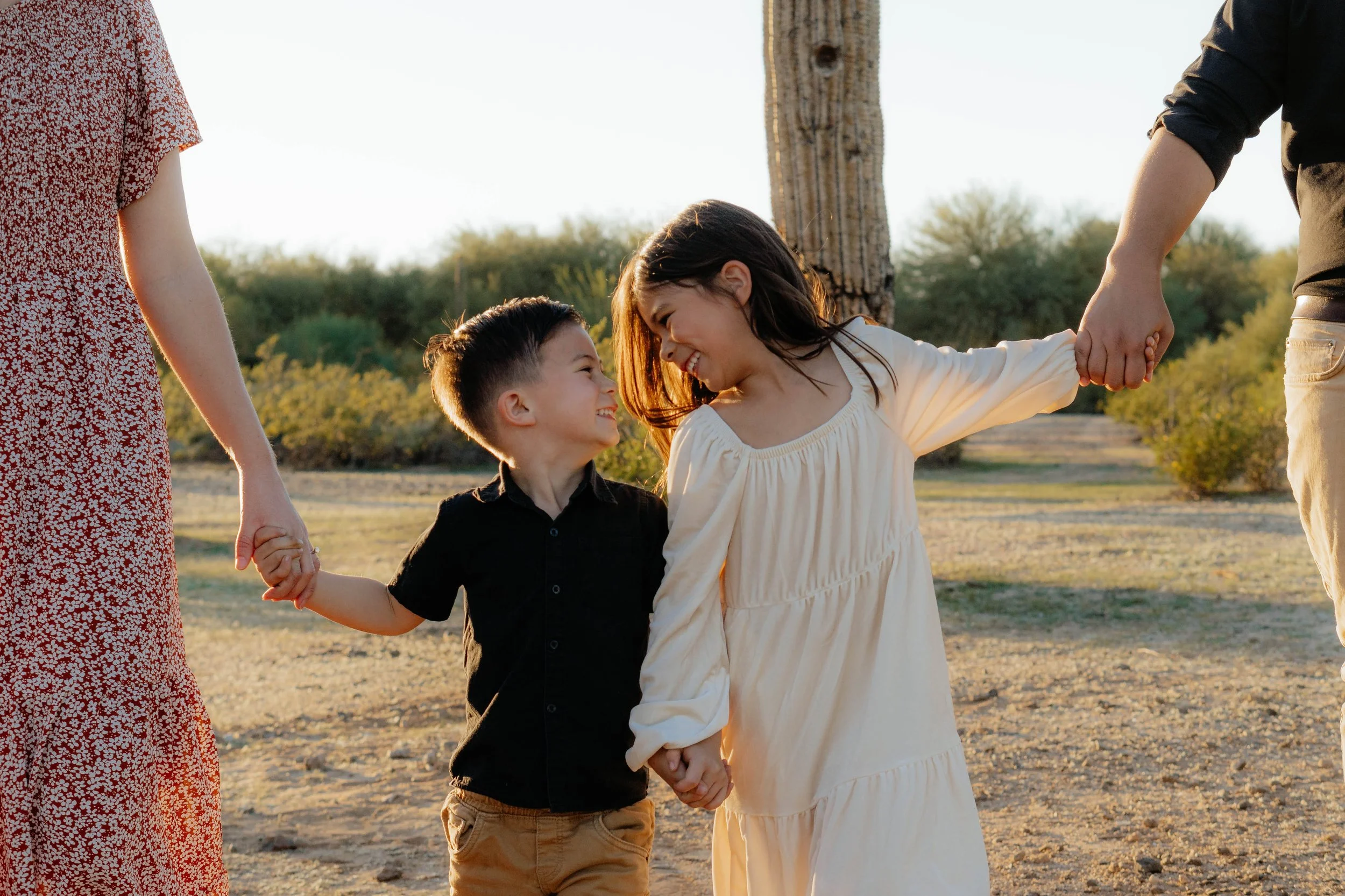 Two children holding hands and smiling, facing each other outdoors during sunset, with parents holding their hands and a cactus in the background.