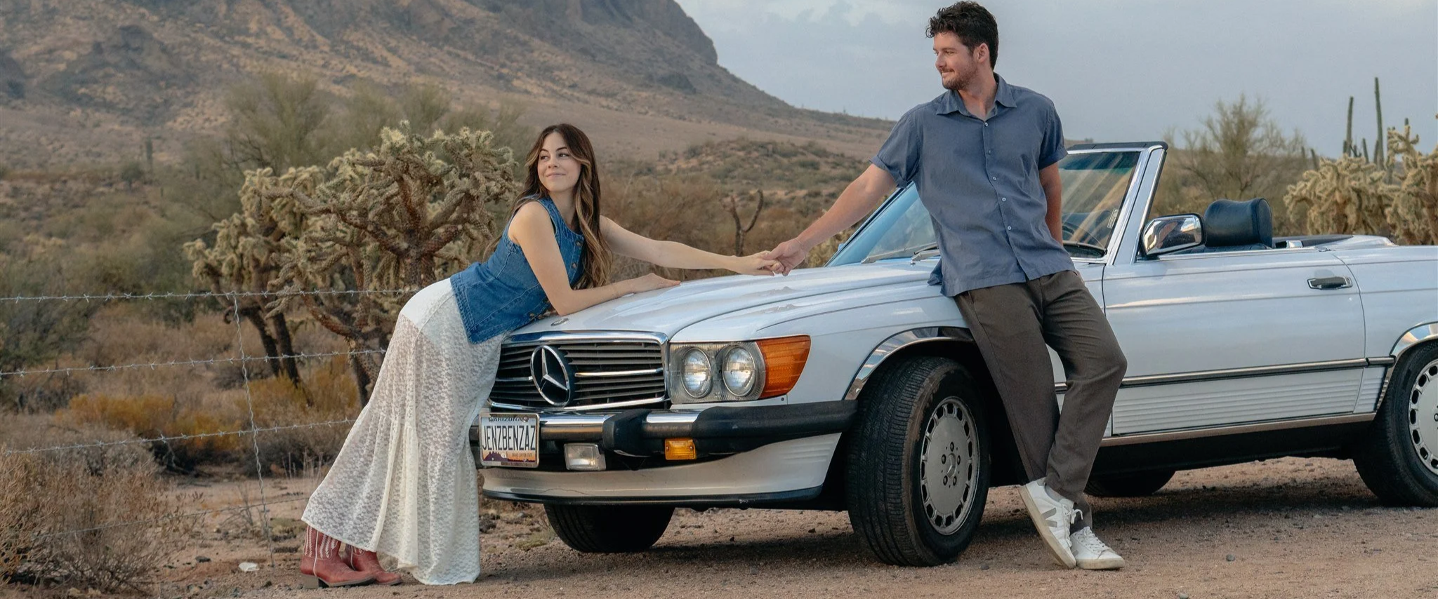 A young woman in a denim jacket and long skirt leans on the front of a silver Mercedes-Benz convertible, holding hands with a man in a blue shirt and gray pants, standing beside the car in a desert landscape with cacti and mountains in the background.