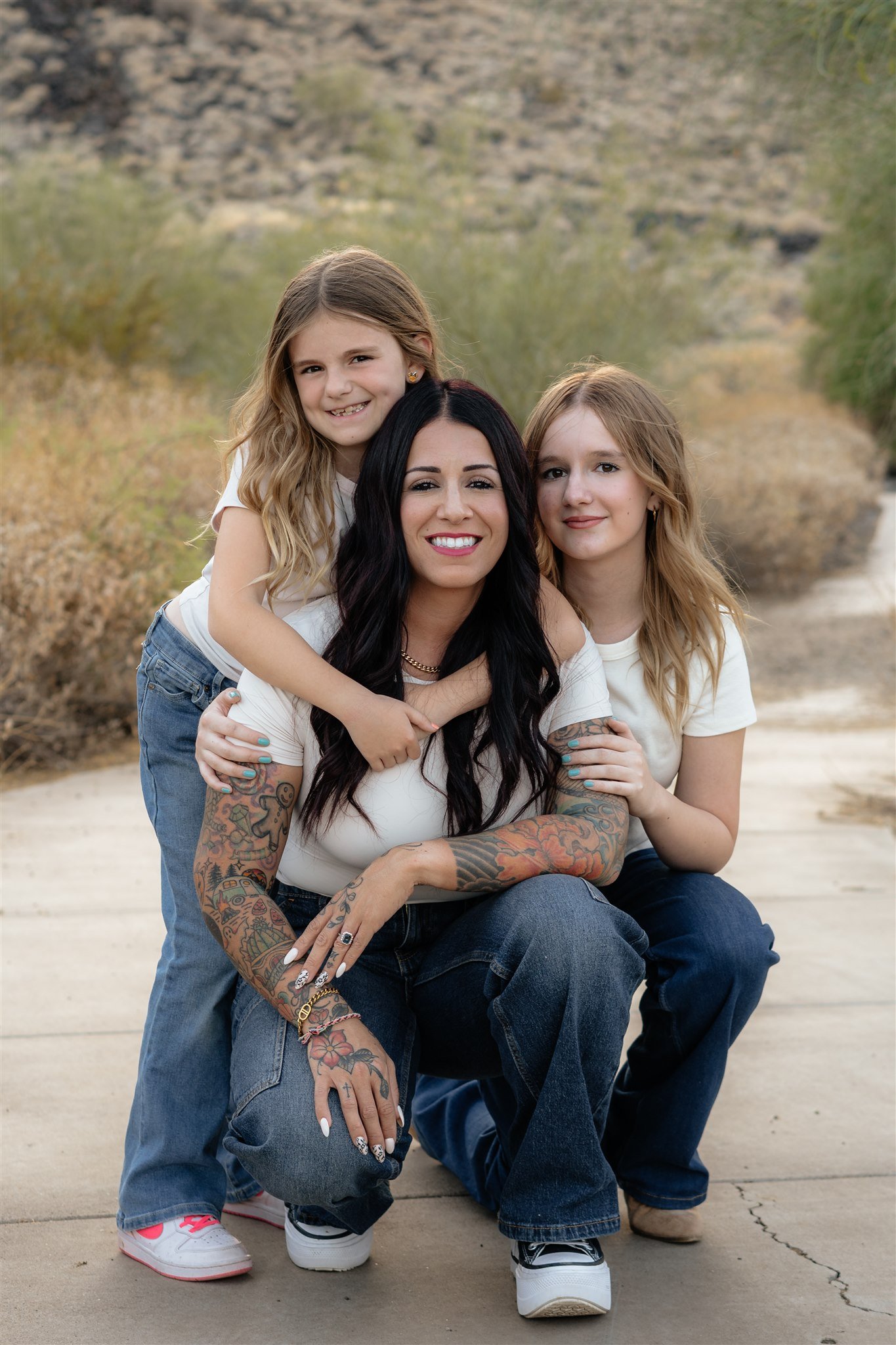 A woman with black hair and tattoos on her arms sitting on the ground with two young girls, one with long blonde hair and the other with light brown hair, outdoors with a blurred desert and mountain background.