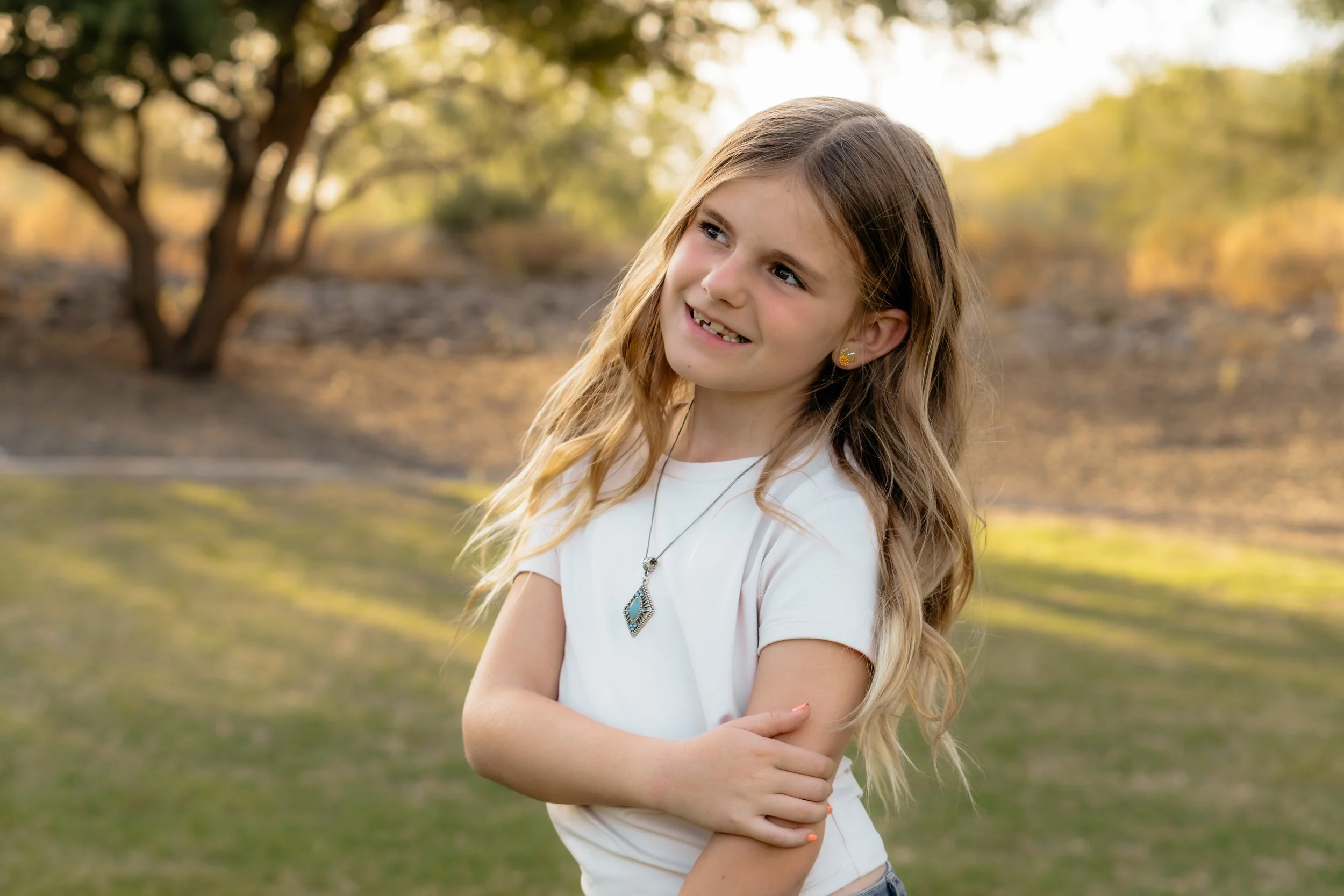 A young girl with long blonde hair, dressed in a white shirt, standing outdoors with trees and greenery in the background, smiling and crossing her arms.