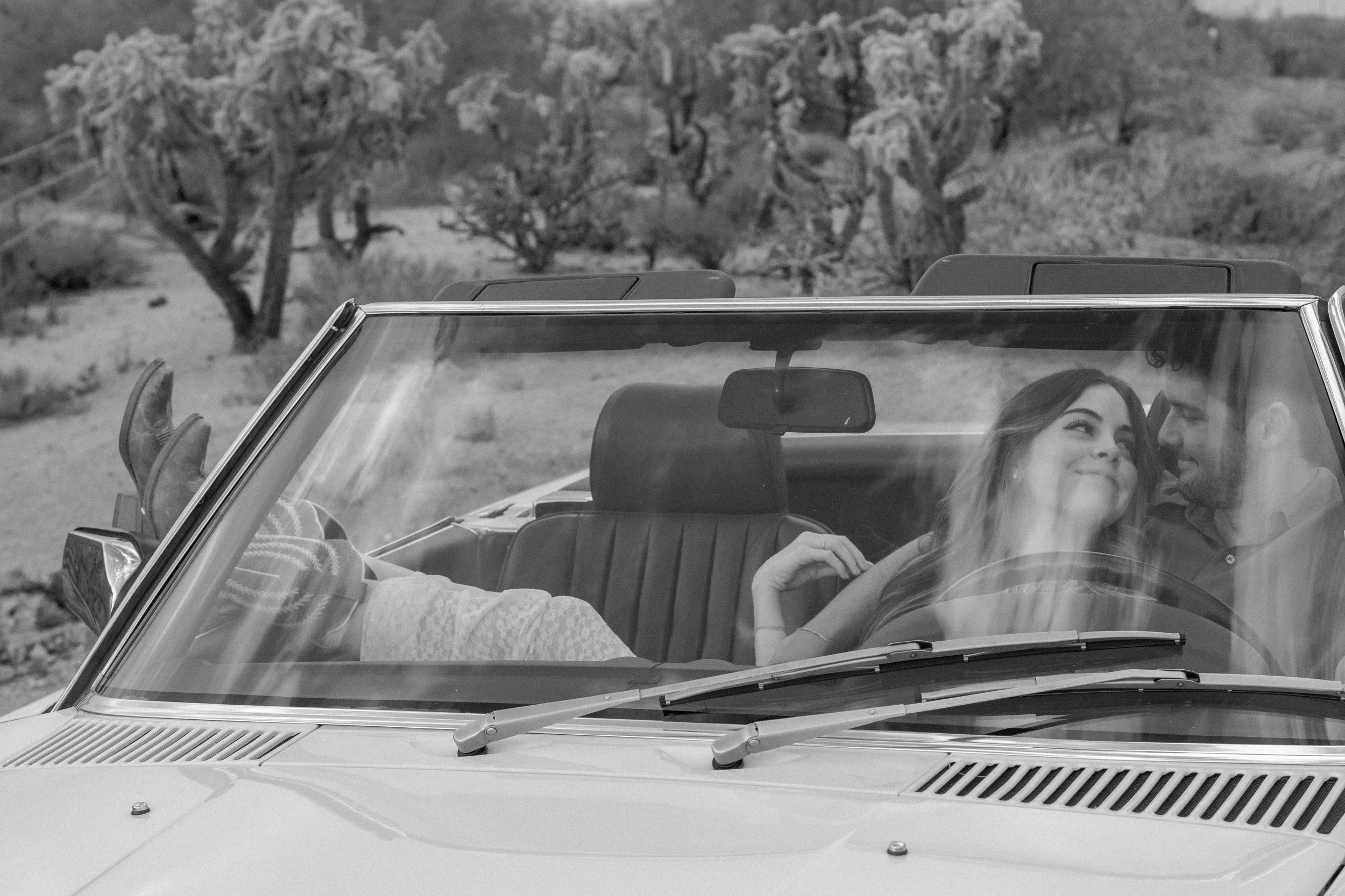 A black-and-white photograph of a man and woman sitting in a convertible car in a desert-like area with sparse vegetation, looking at each other and smiling.