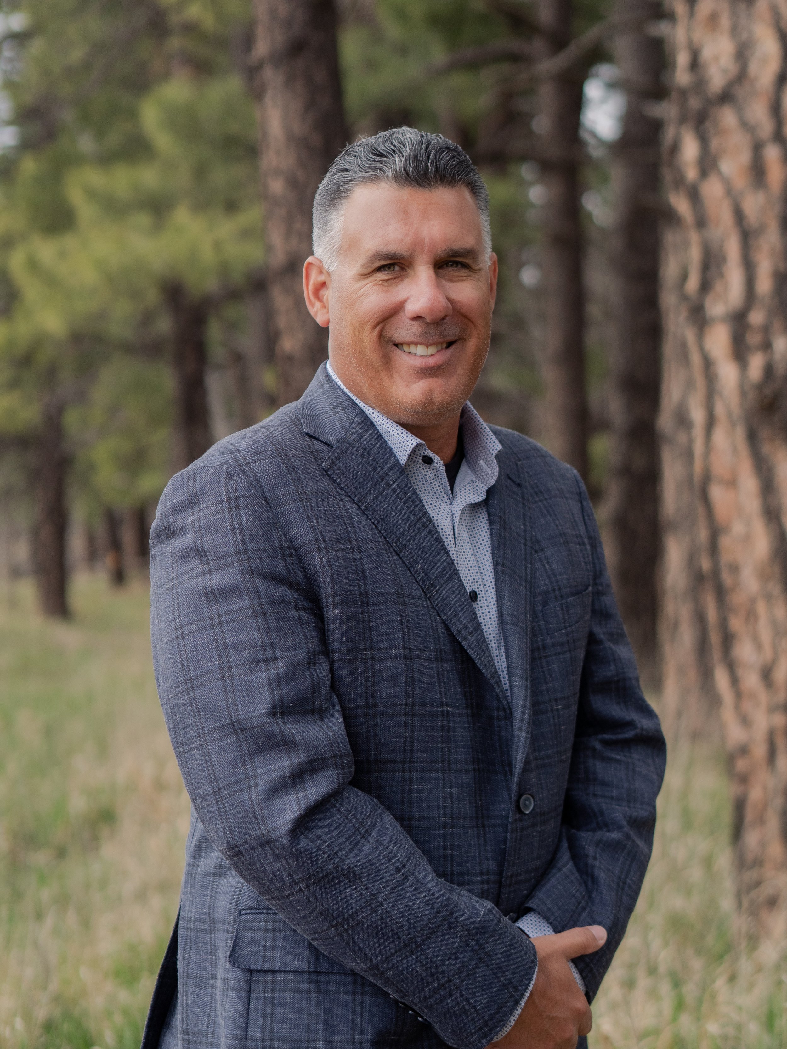 A man in a plaid suit smiling outdoors with trees in the background.