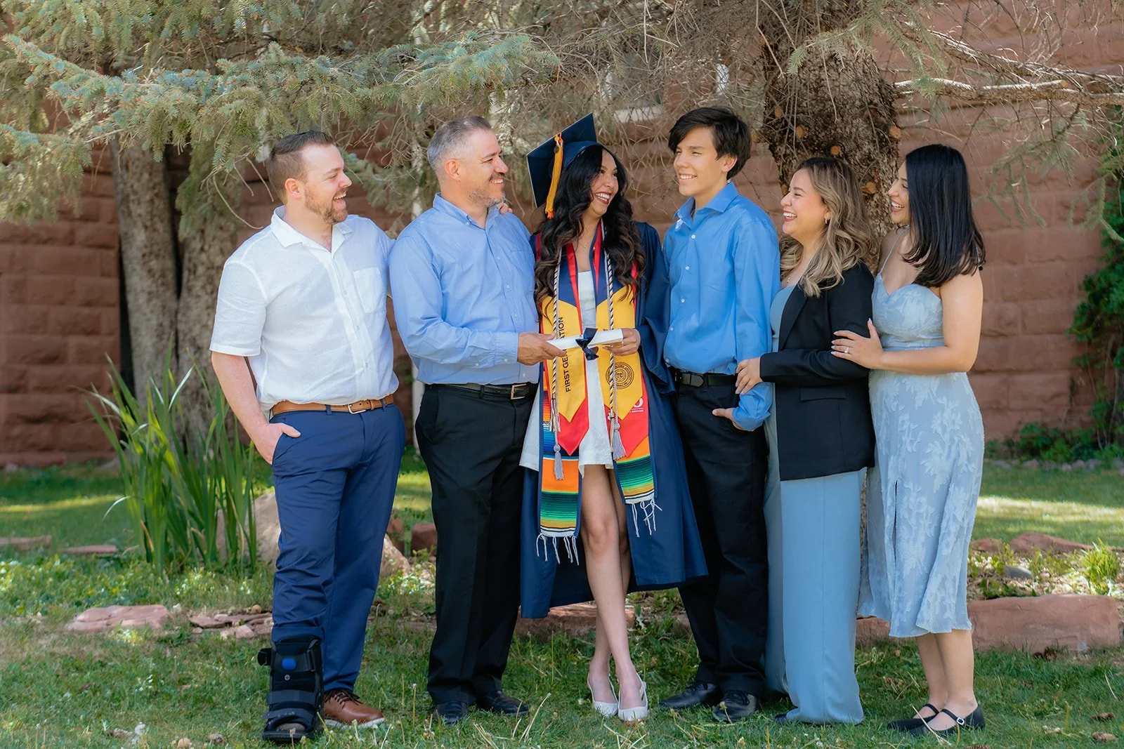 A woman in graduation gown and cap holding diploma, surrounded by five people smiling outdoors, with trees and a brick wall in the background.