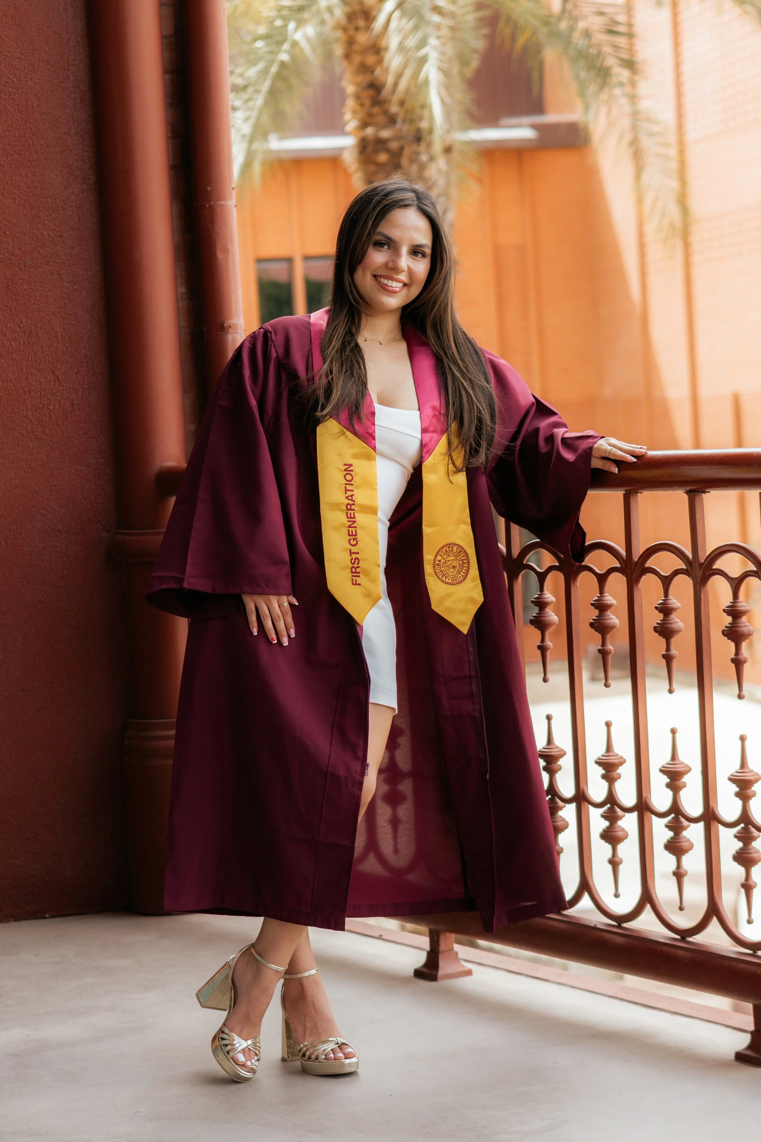 A young woman in a maroon graduation gown and yellow stole, standing on a balcony outside of a building with palm trees in the background, smiling, wearing high heels.