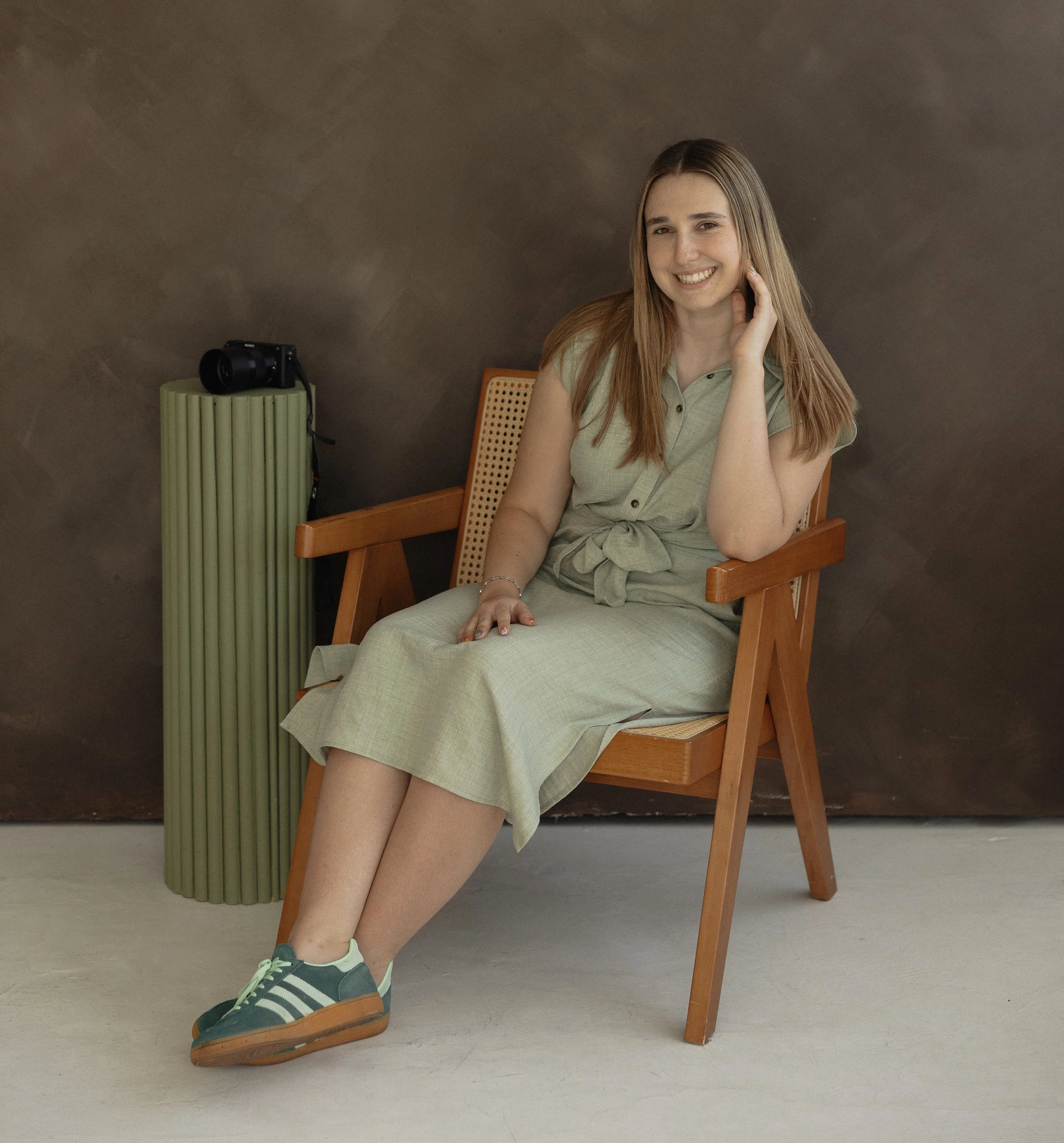 A young woman with long, light brown hair sitting on a wooden chair with cane backrest, smiling and touching her hair with her right hand, wearing a light green dress and green sneakers with white laces, in front of a dark brown wall with a lime green vintage-looking camera placed on a green cylindrical side table to her left.
