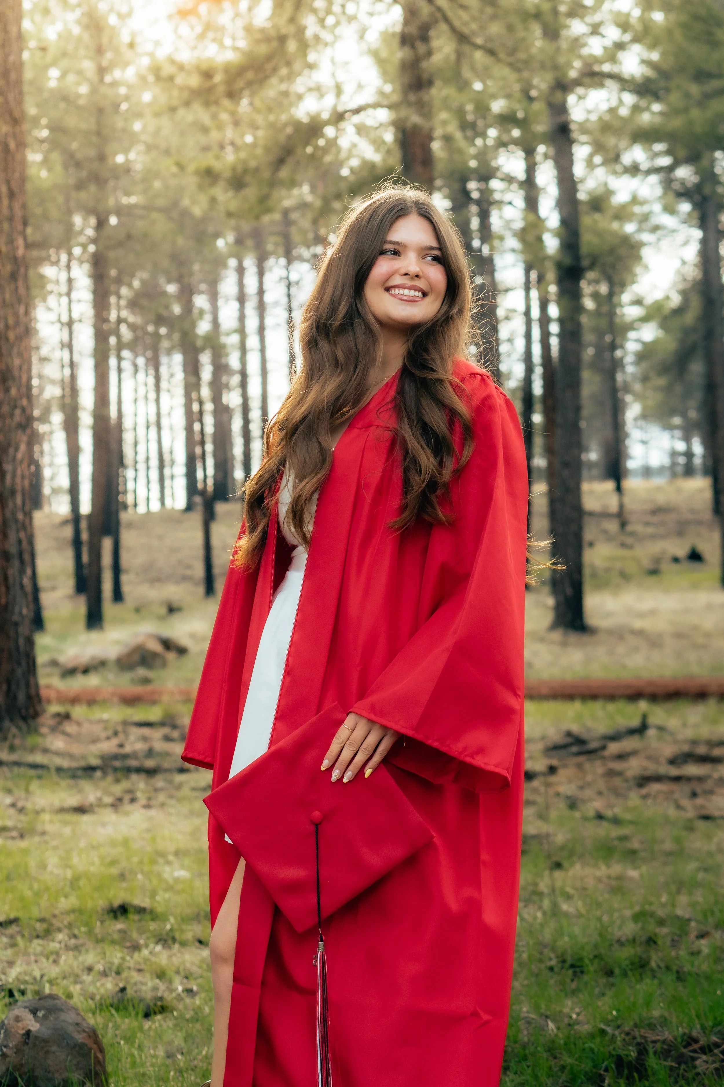 A young woman in a red graduation gown and cap stands outdoors in a forest area with tall trees and sunlight filtering through the branches, smiling and looking to her right.