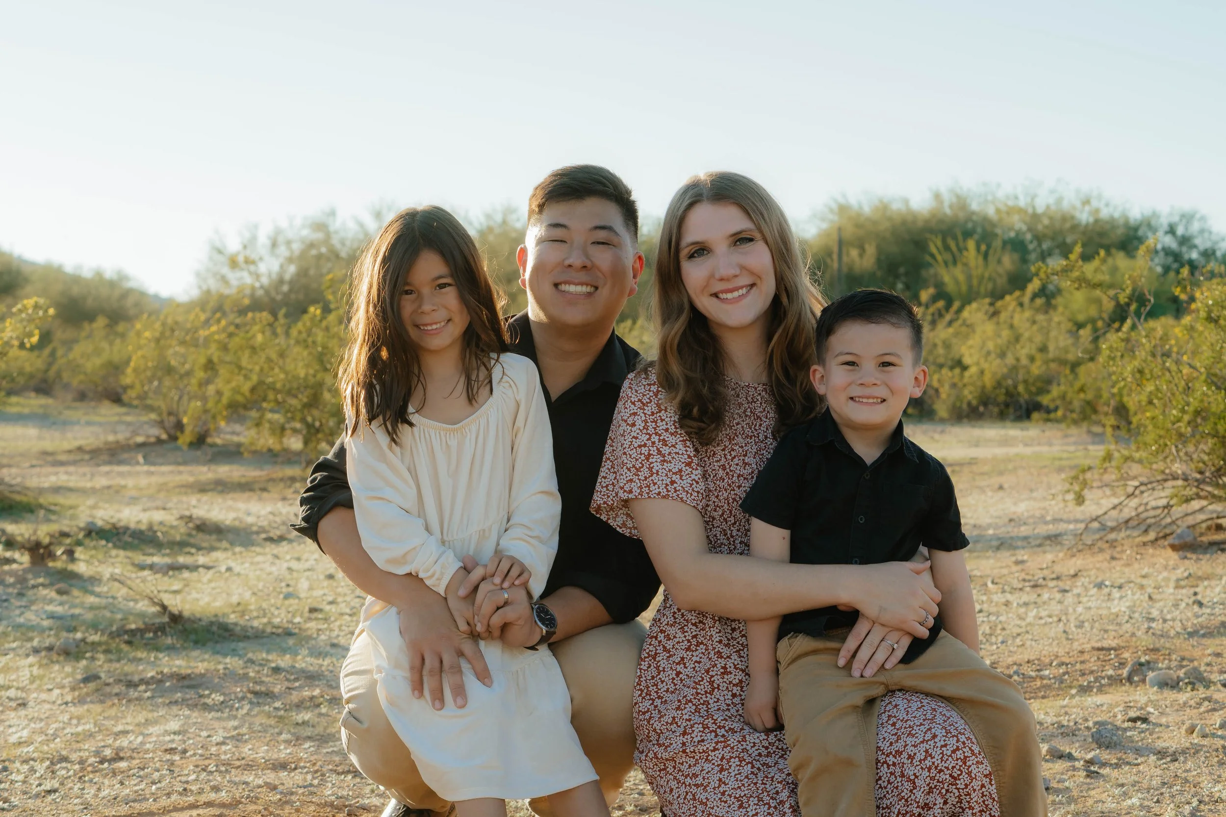 A family of four smiling outdoors in a natural setting with green trees in the background.