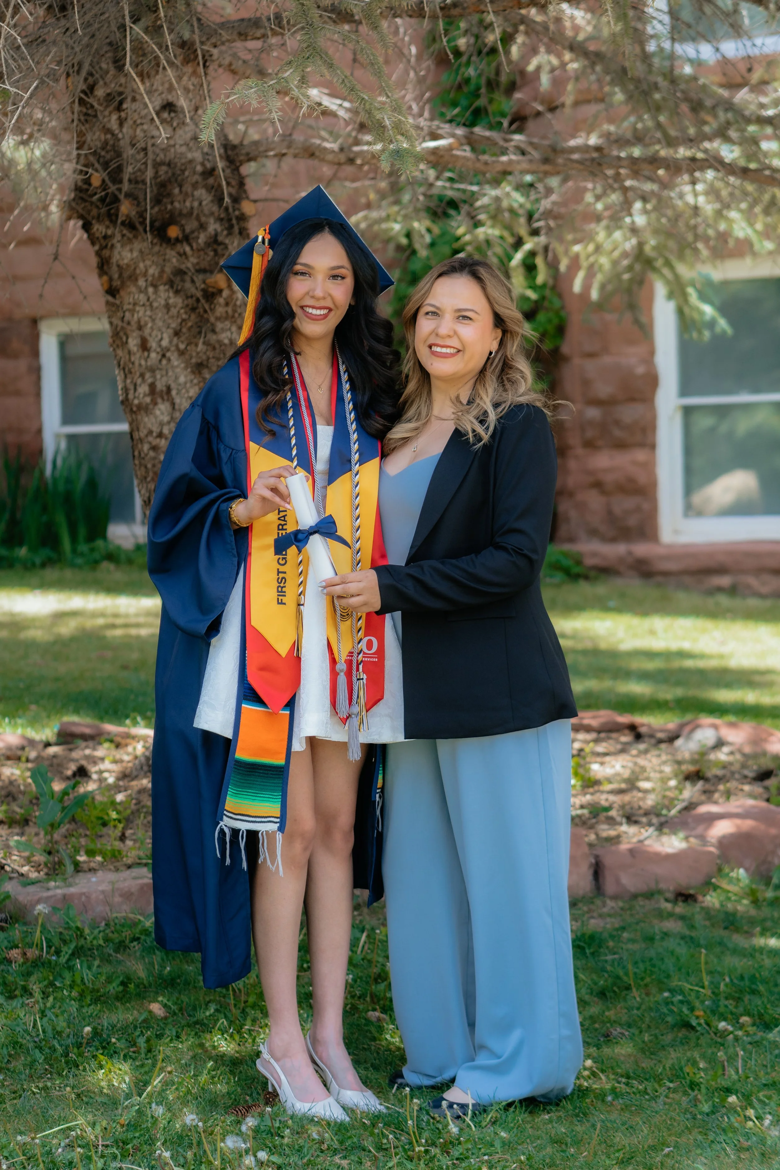 A young woman in a graduation gown and cap holding a diploma, smiling, with a woman in formal attire standing beside her, outdoors near a tree and a brick building.