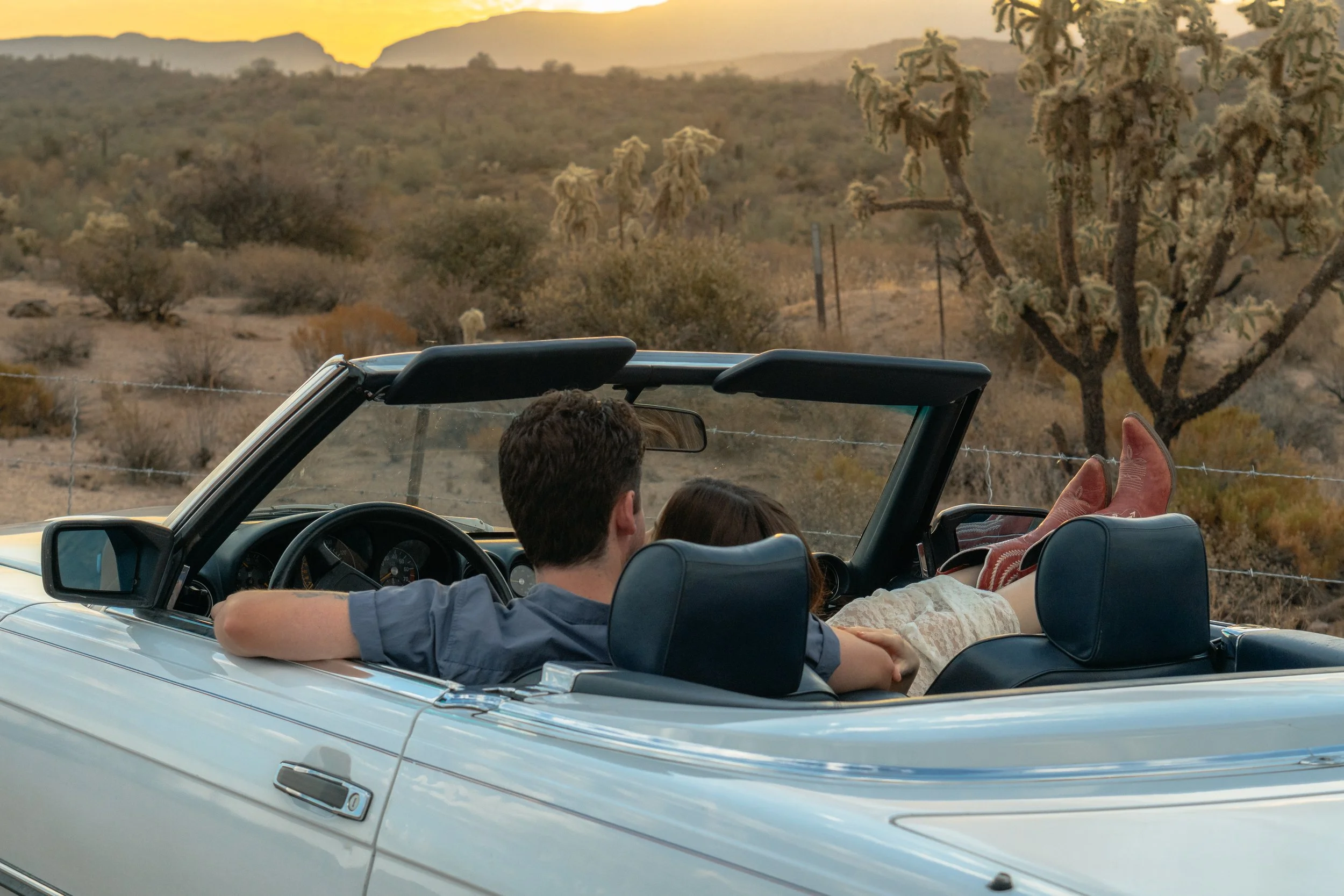 Two people in a white convertible car relaxing in a desert landscape during sunset, with cacti and mountains in the background.
