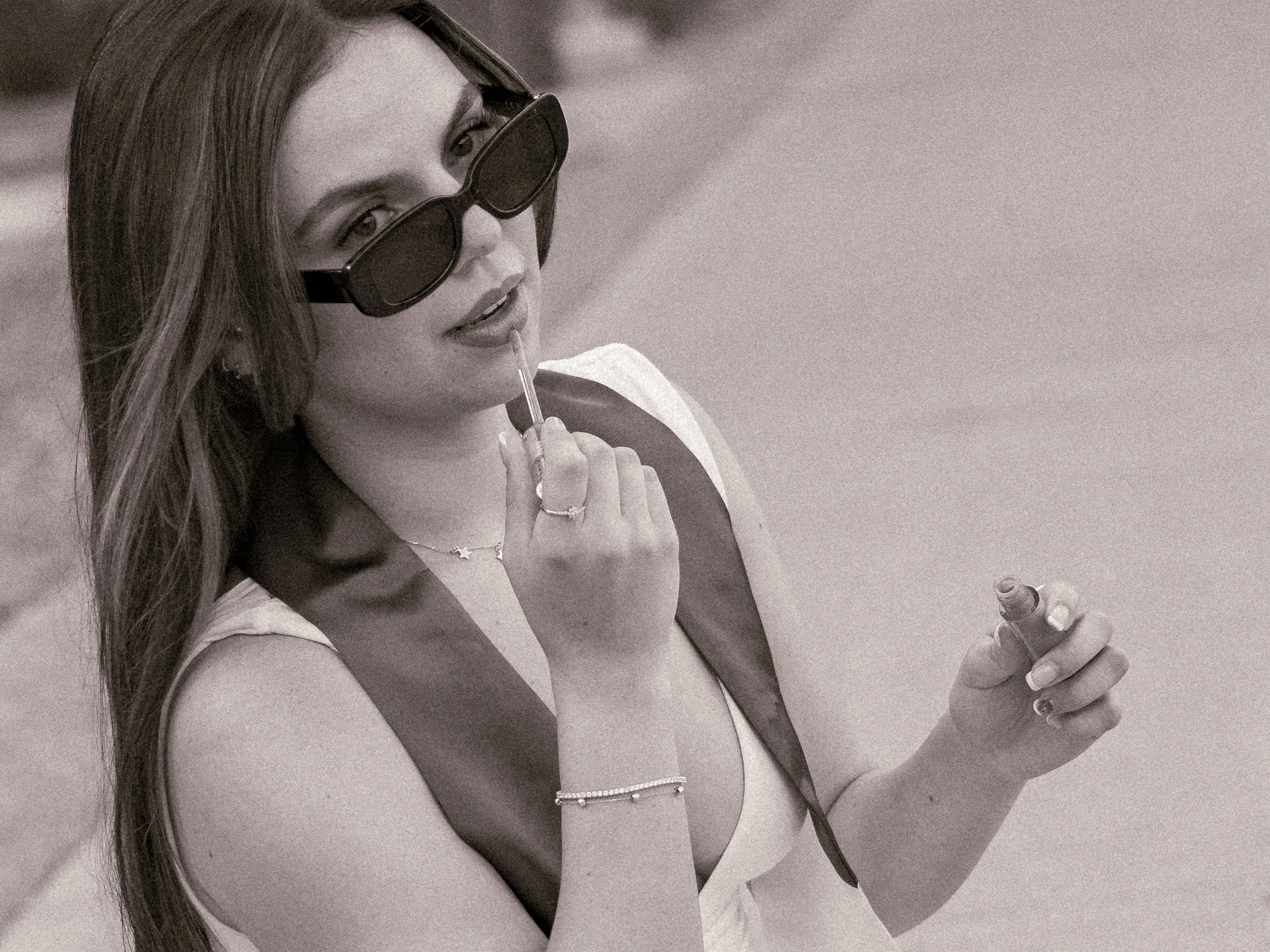 A woman with long hair wearing sunglasses, holding lipstick, and wearing layered jewelry including rings, necklaces, and bracelets.
