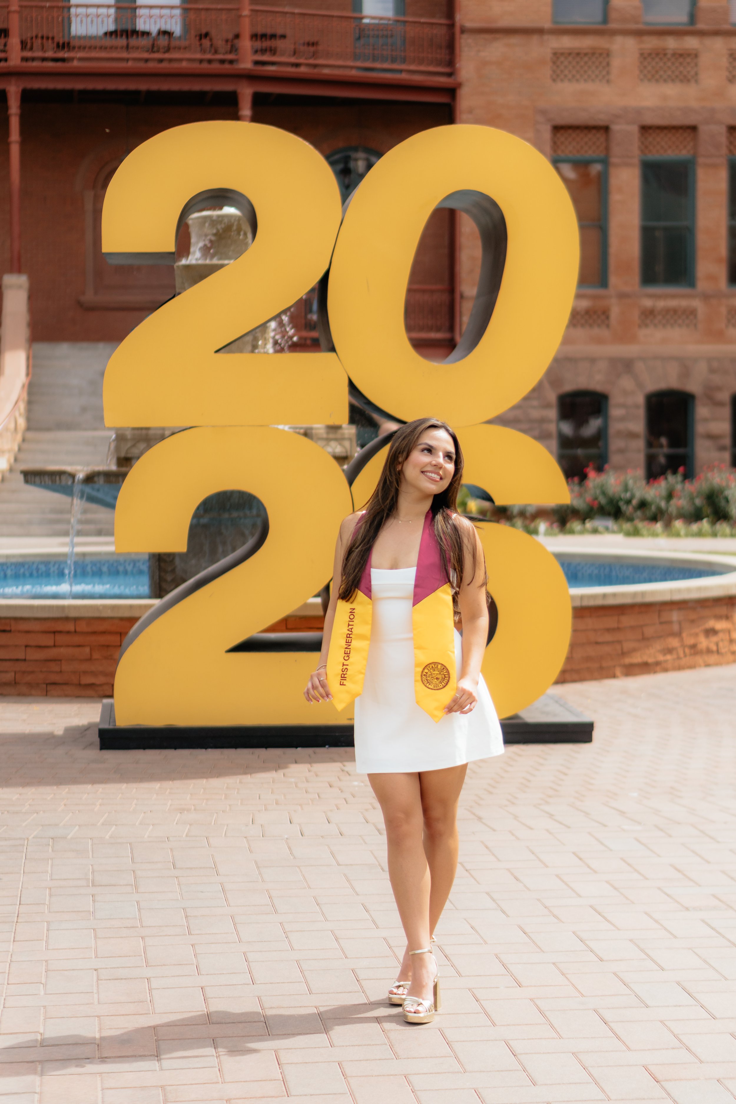 A young woman in a white dress and high heels stands in front of a large yellow "2026" sign with a fountain behind it. The woman is smiling and wears a yellow sash that says "First Generation".