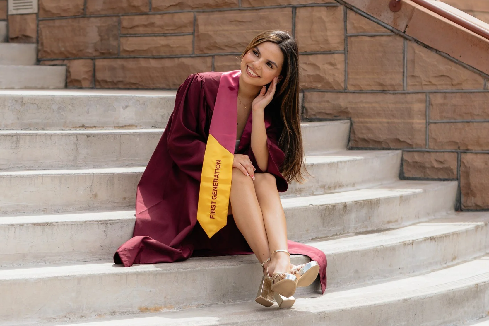 A young woman in a maroon graduation gown with a yellow stole that reads 'First Generation' sitting on outdoor stairs, smiling, with one hand resting on her face and her legs crossed, wearing tan high-heeled sandals.