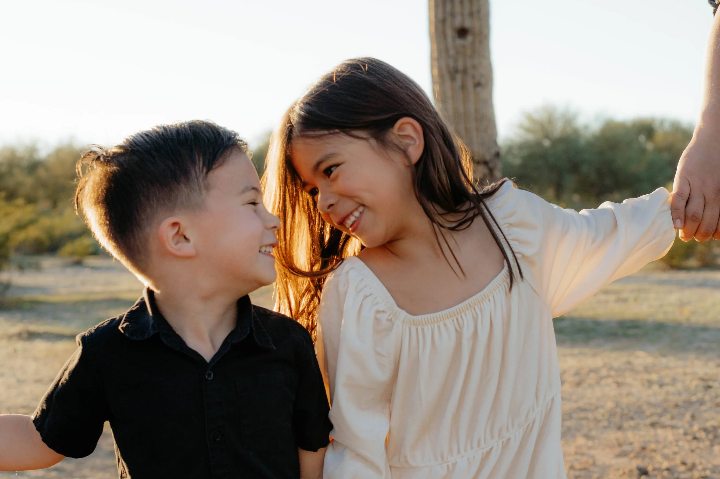 A boy and a girl smiling and looking at each other outdoors during sunset, with the girl lifting her arm.