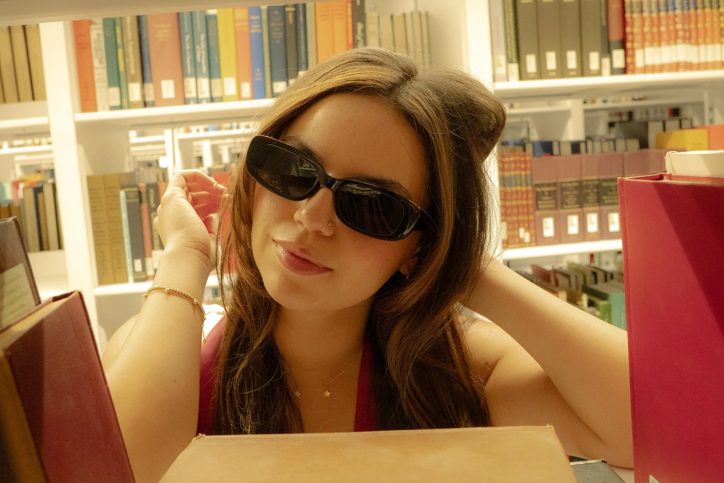 A woman wearing sunglasses, a nose piercing, and a star necklace, sitting at a table in a library surrounded by colorful books.