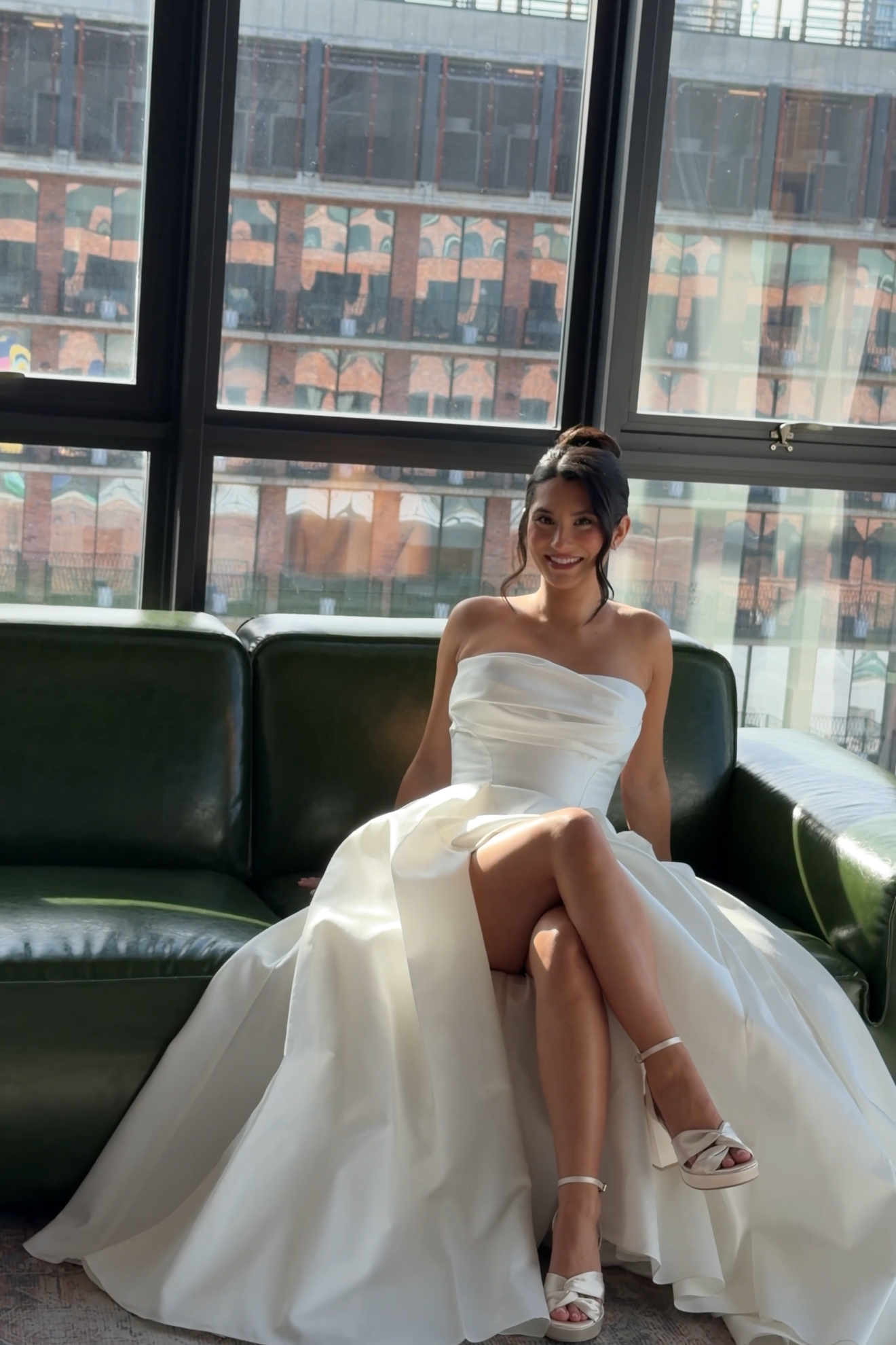 A woman in a white dress sitting on a black leather sofa, smiling with a cityscape of a brick building with large windows in the background.