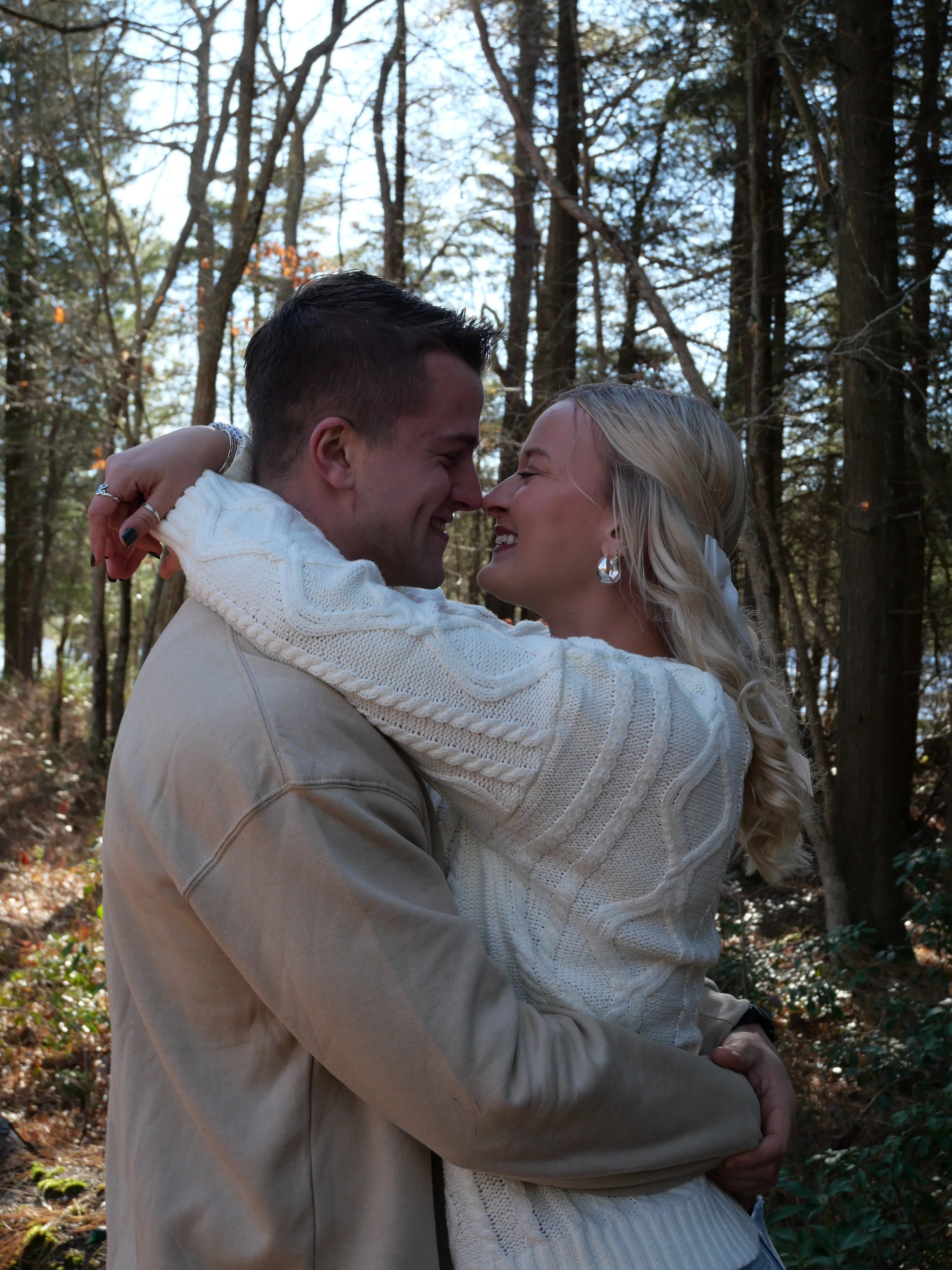 A happy couple embraces in a forested outdoor setting, with trees and sunlight in the background.