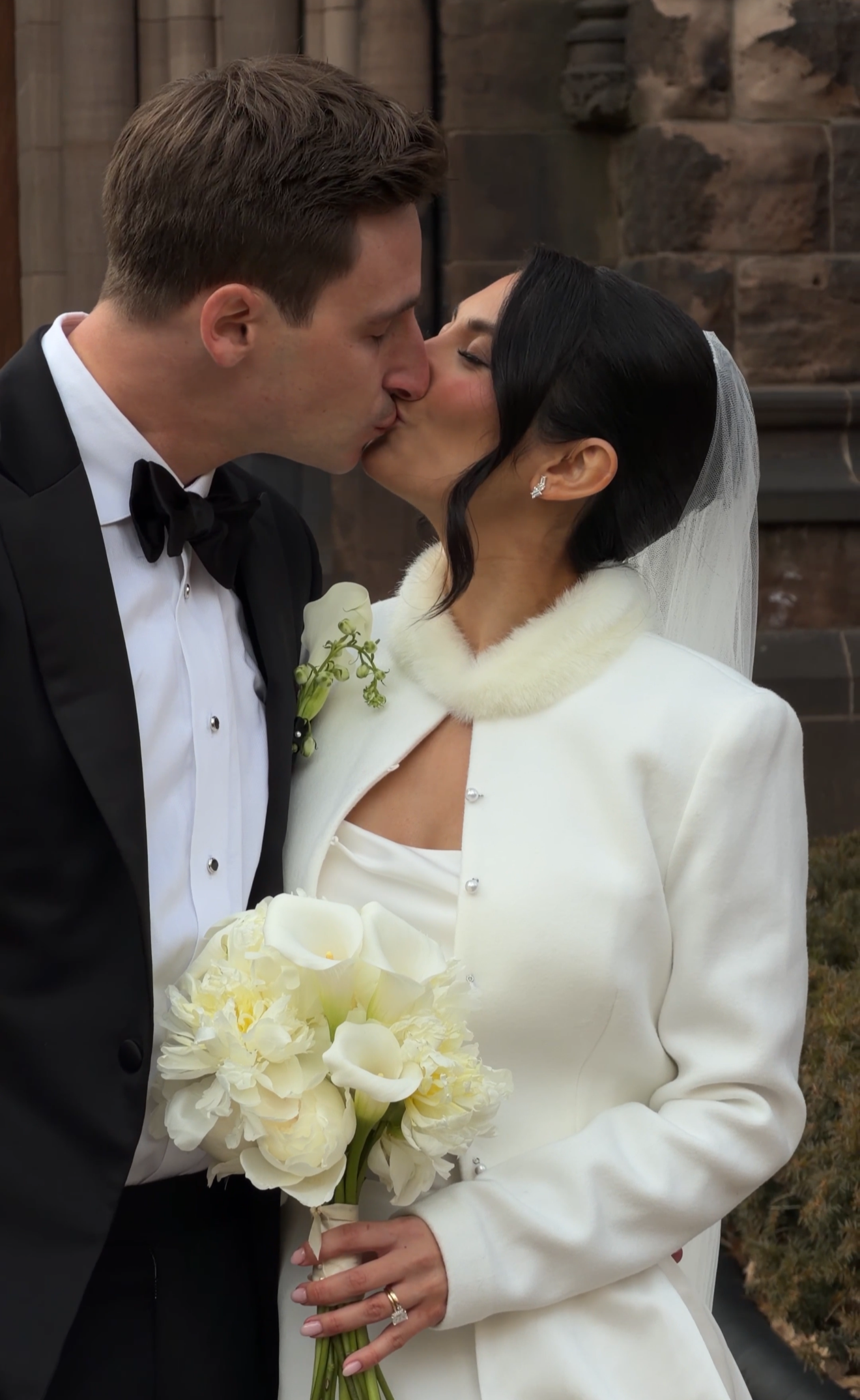 A bride and groom share a kiss during their wedding ceremony, with the bride holding a bouquet of white calla lilies and wearing a white coat with a fur collar.