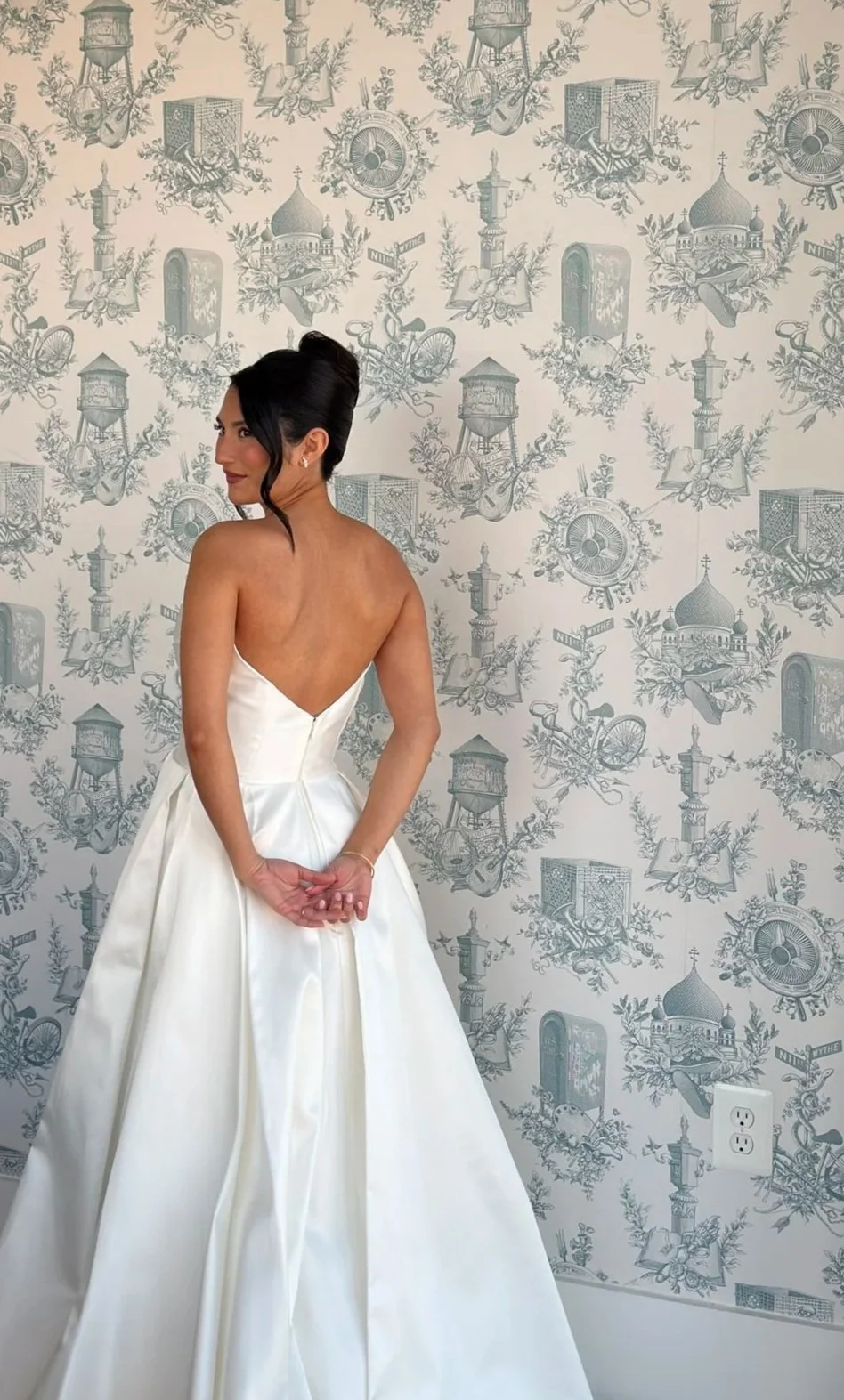 A woman in a strapless white wedding dress standing in front of a patterned wallpaper with illustrations of landmarks, bicycles, and architectural elements, posing with her hands clasped behind her back and looking over her shoulder.