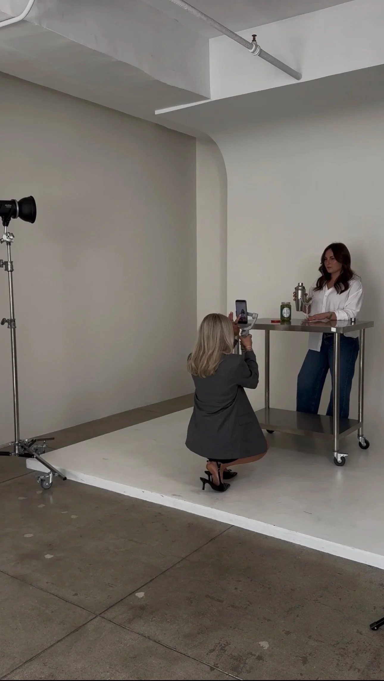 A woman in a white blouse and jeans standing behind a metal cart with drinkware and a can on it, being photographed by another woman kneeling on a white photography floor mat, with a lighting setup to the left, in an indoor studio with plain walls.