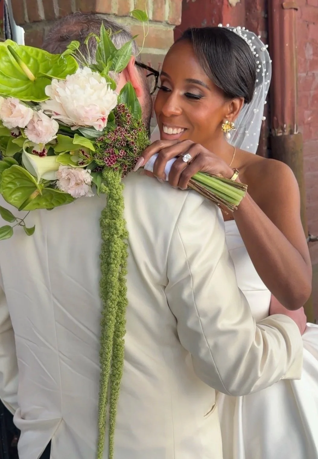 A bride and groom are hugging, with the bride smiling and holding a bouquet of flowers. The bride is wearing a veil with pearl details, earrings, and a ring, and has her eyes closed in a happy expression. The groom is dressed in a cream-colored suit, and their embrace is intimate and joyful.
